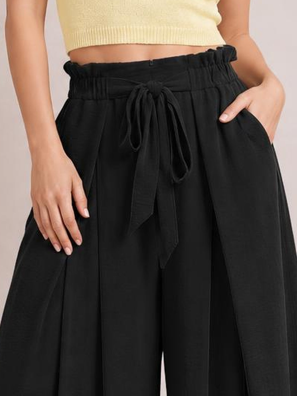 A model wears black tied wide leg pants with a gathered elastic waistband, long drawstring, relaxed silhouette, and side pockets, posing with one hand tucked inside.