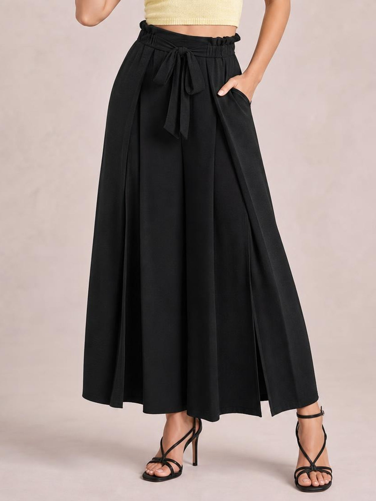 Model wears black tied wide-leg pants with a high gathered waist, flowing full-length silhouette, side slit detail, and a hand in the pocket.