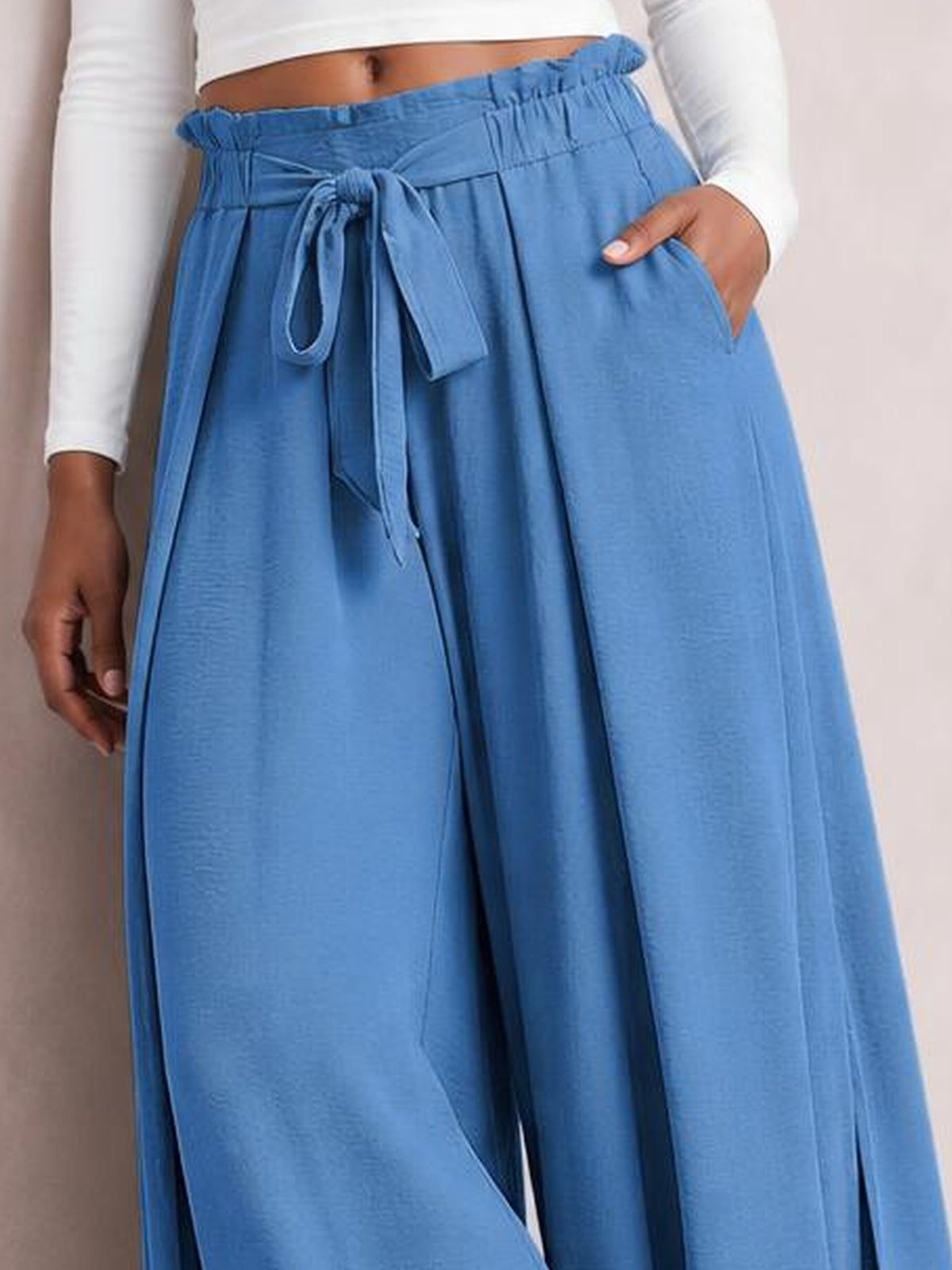 A cropped model wears blue tied slit wide-leg pants with an elastic waist, flowing silhouette, front drawstring, and side pockets, resting one hand inside a pocket.