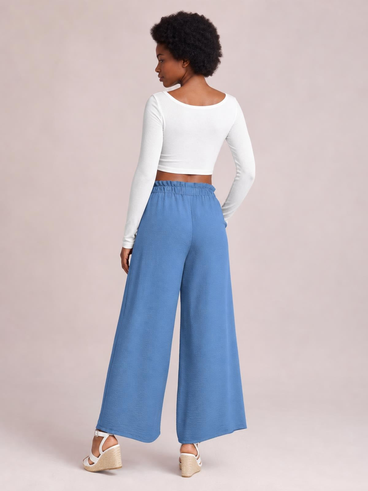 Model turned away in blue tied slit wide leg pants with an elastic waist and flowing full-length silhouette, paired with a white long-sleeve crop top.