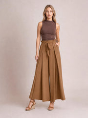 Model wearing caramel-brown tied slit wide-leg pants with a high gathered waistband, flowing floor-length silhouette, and relaxed pose with one hand in a pocket.