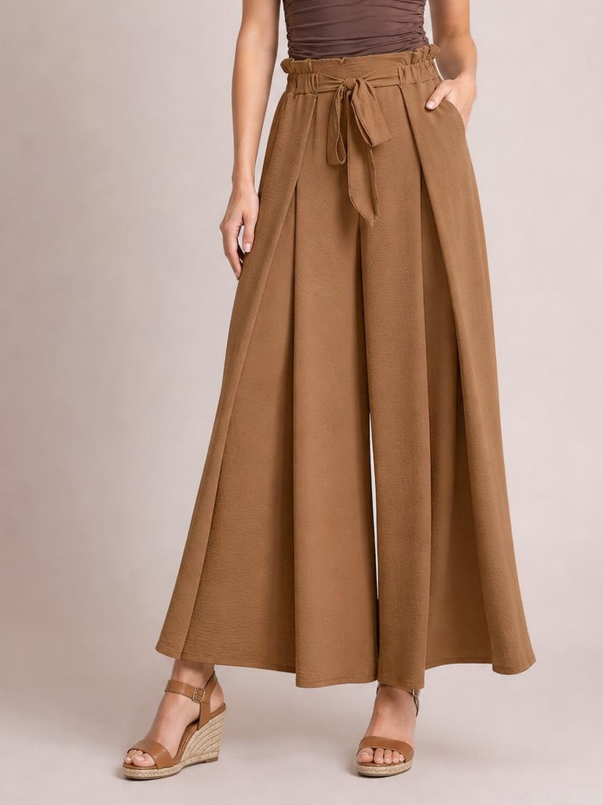 A model wears brown tied wide-leg pants with a gathered high-rise waist, front sash, flowing full-length silhouette, and hands resting in the pockets.