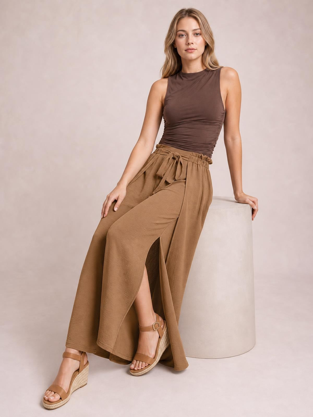 Model seated in tan tied slit wide-leg pants, featuring a relaxed flowing silhouette, gathered waistband, and dramatic front leg openings.