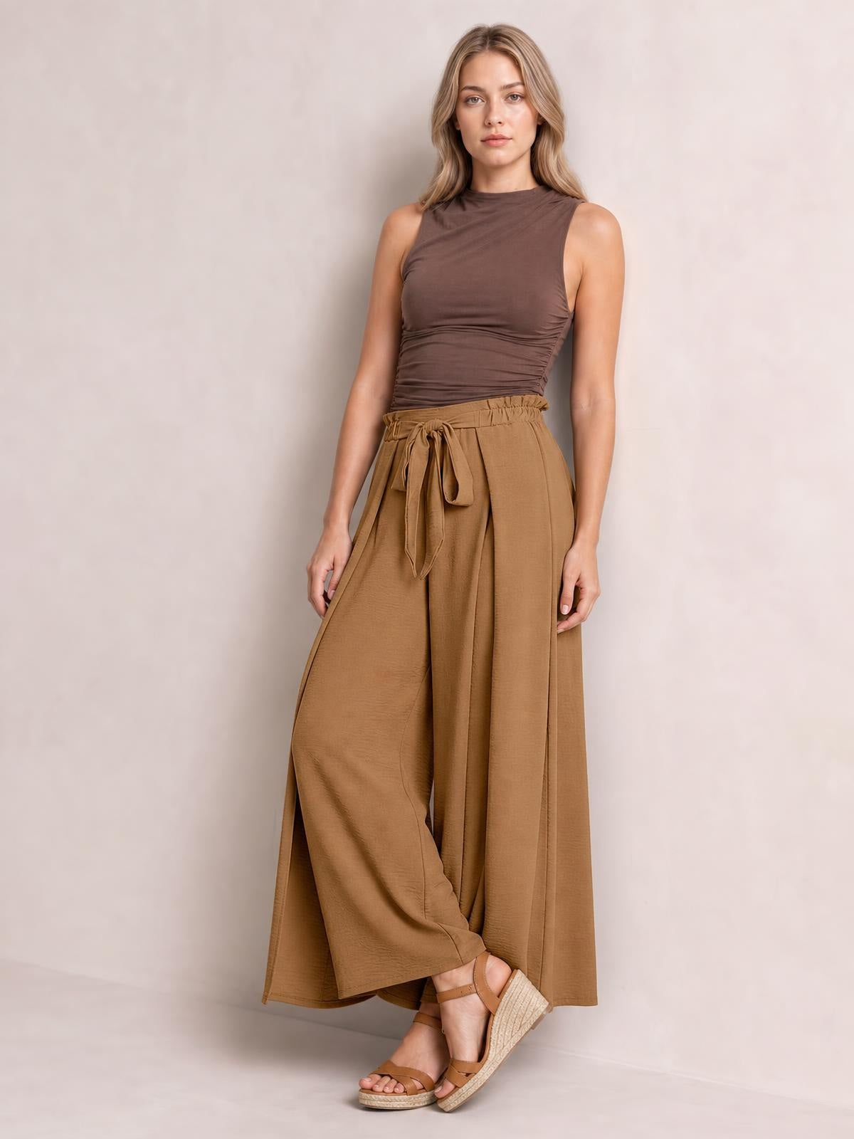 Model poses in brown tied slit wide-leg pants with a high gathered waist, flowing opaque fabric, and a floor-length wide silhouette.