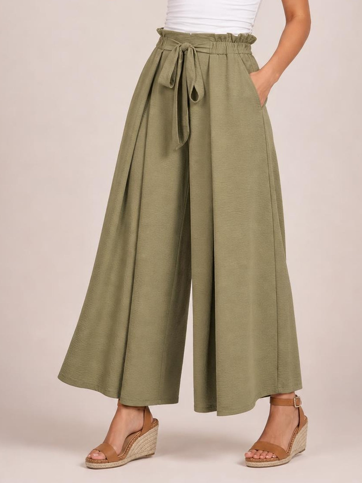 A model wears olive-green tied slit wide-leg pants with a gathered waist, flowing cropped silhouette, and hands-in-pockets pose.
