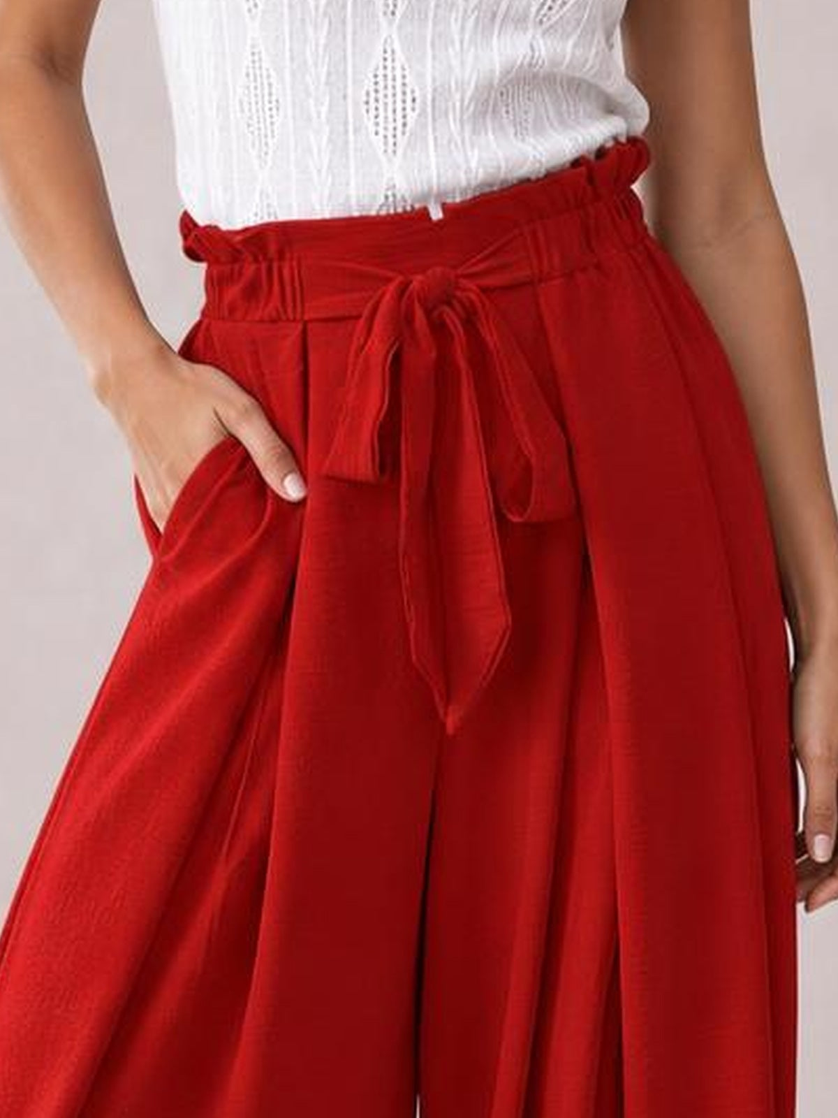 Model wears red opaque wide-leg pants with a tied paperbag waist, flowing silhouette, and a visible side slit while resting one hand in the pocket.