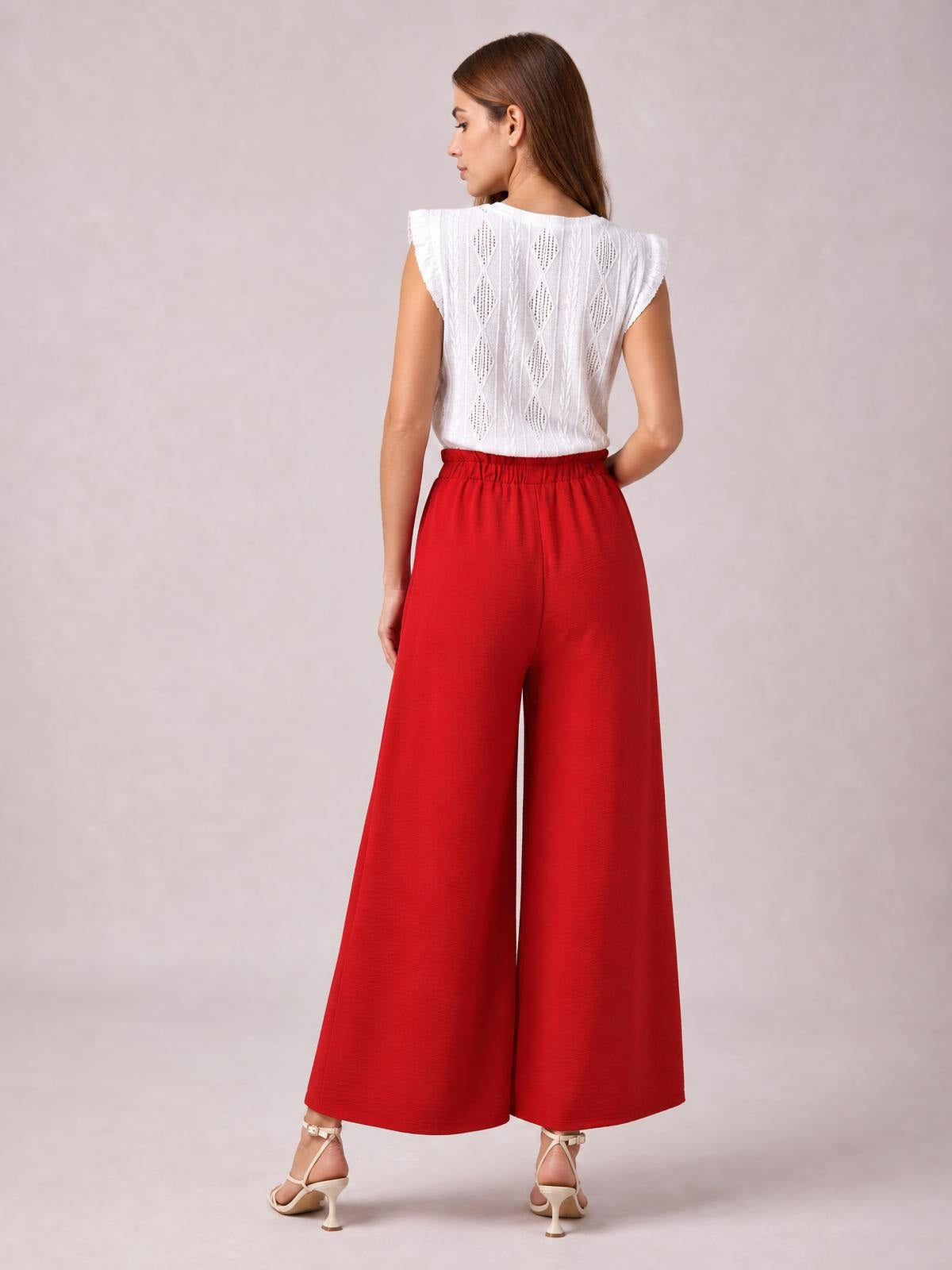 A model poses from behind in vibrant red wide-leg pants with a gathered waistband and flowing full-length silhouette, one hand resting at her waist.