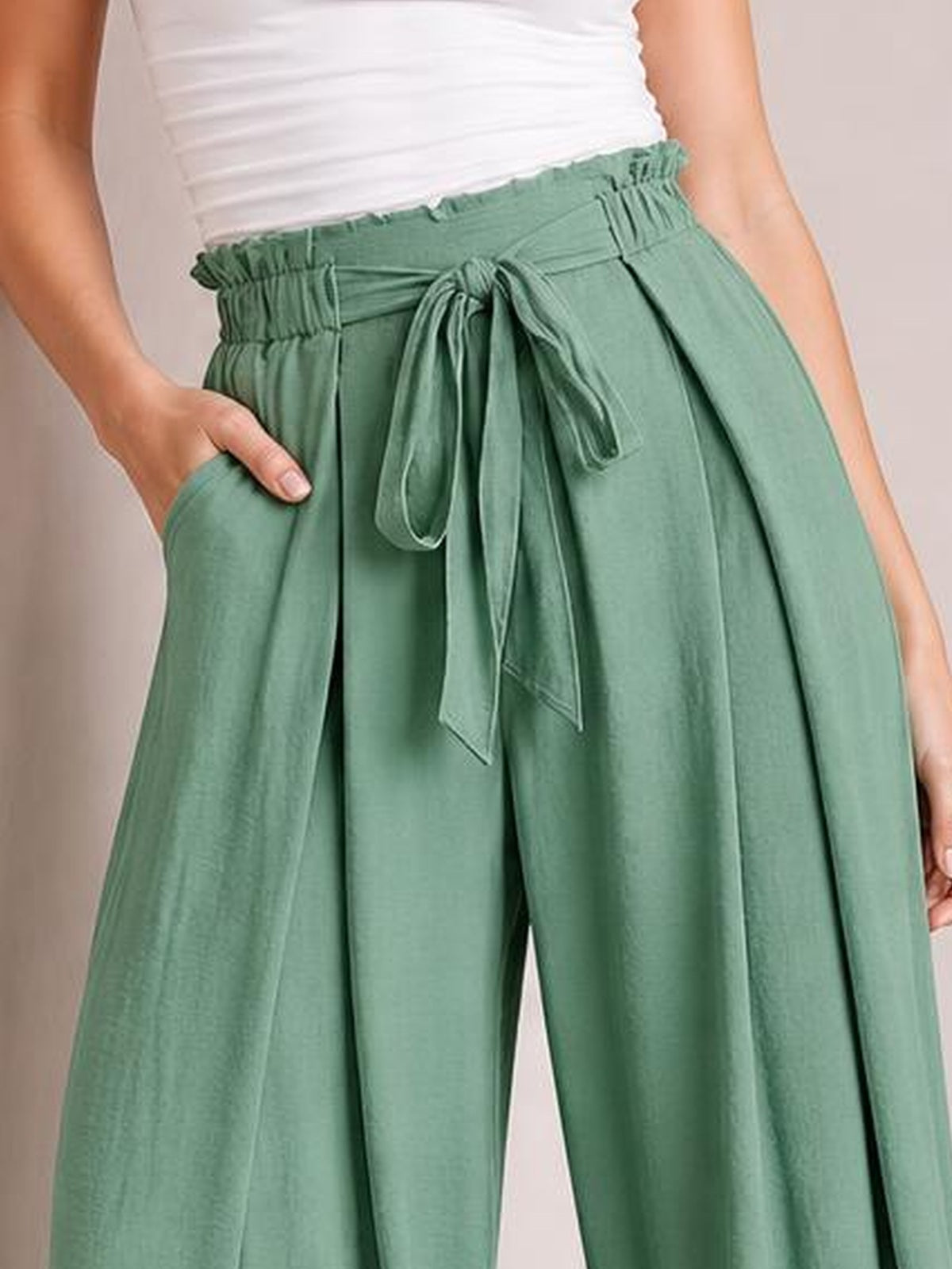 Model wearing sage green tied slit wide leg pants with a high elasticized waist, flowing wide-leg silhouette, and one hand resting in a pocket.