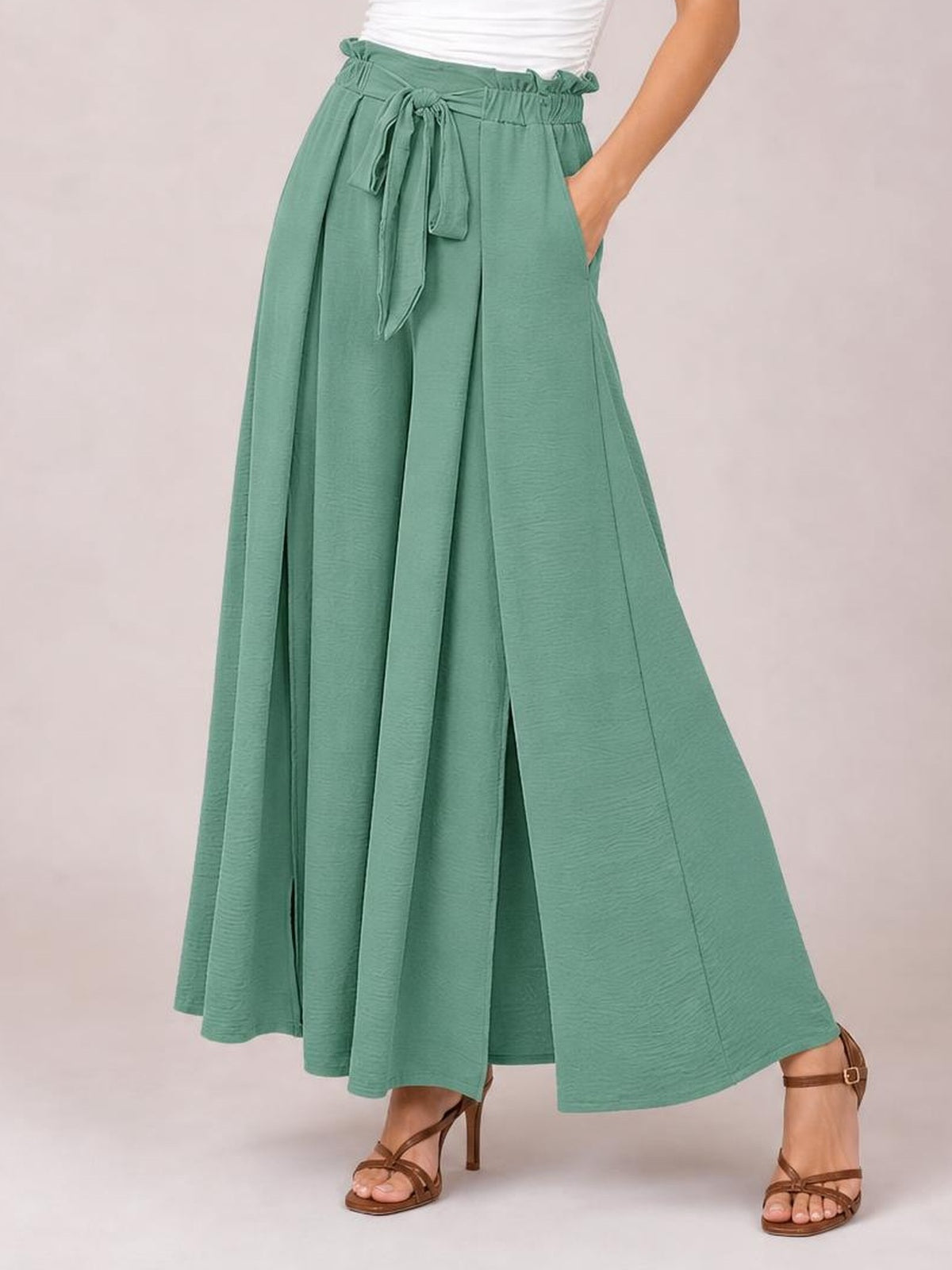A model wears sage green tied slit wide-leg pants with a gathered waist, front sash, flowing silhouette, and hands tucked into the side pockets.