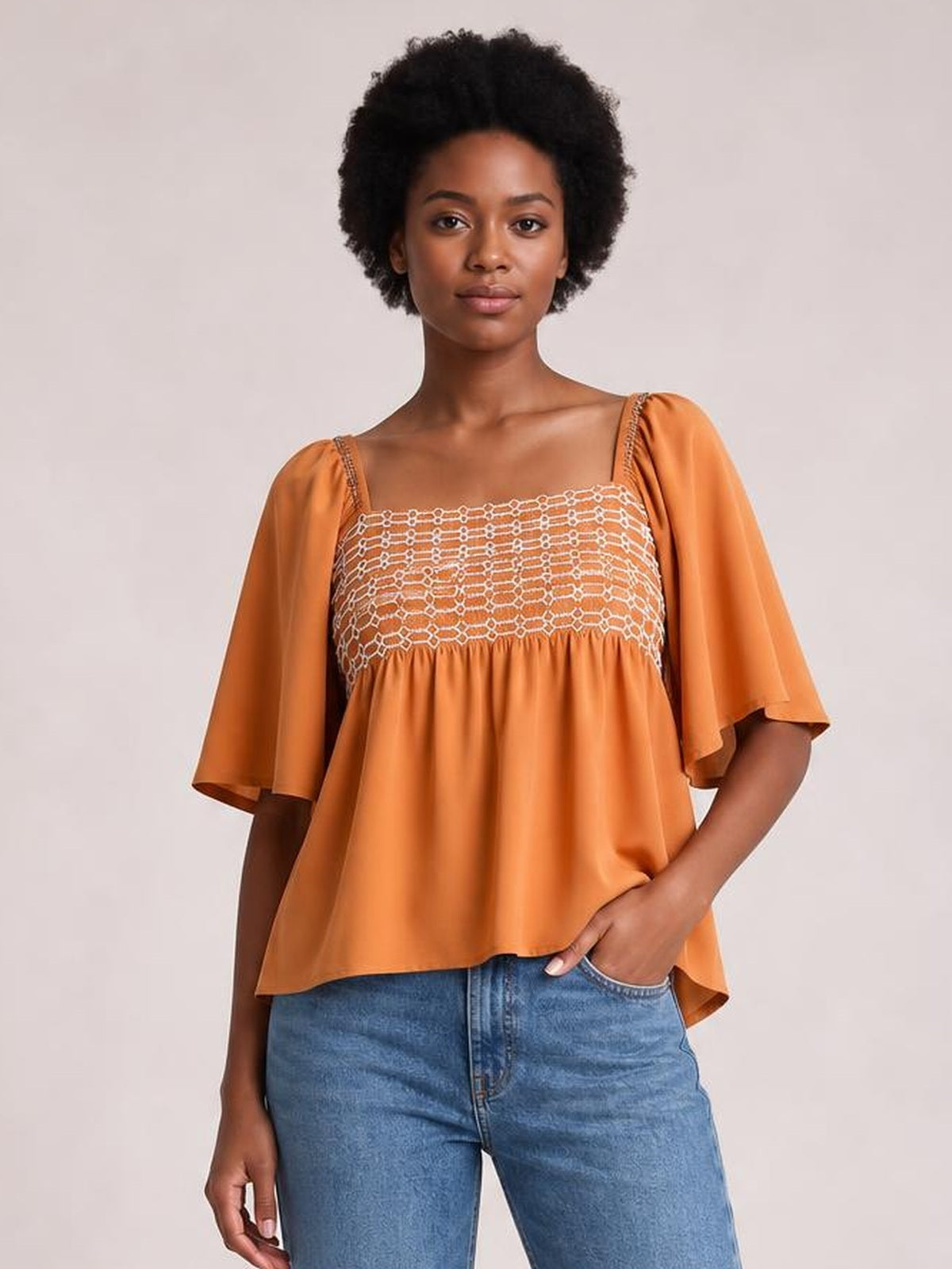 A model wears an orange tied square-neck half-sleeve blouse with a smocked yoke, gathered waist, and relaxed flowing silhouette.