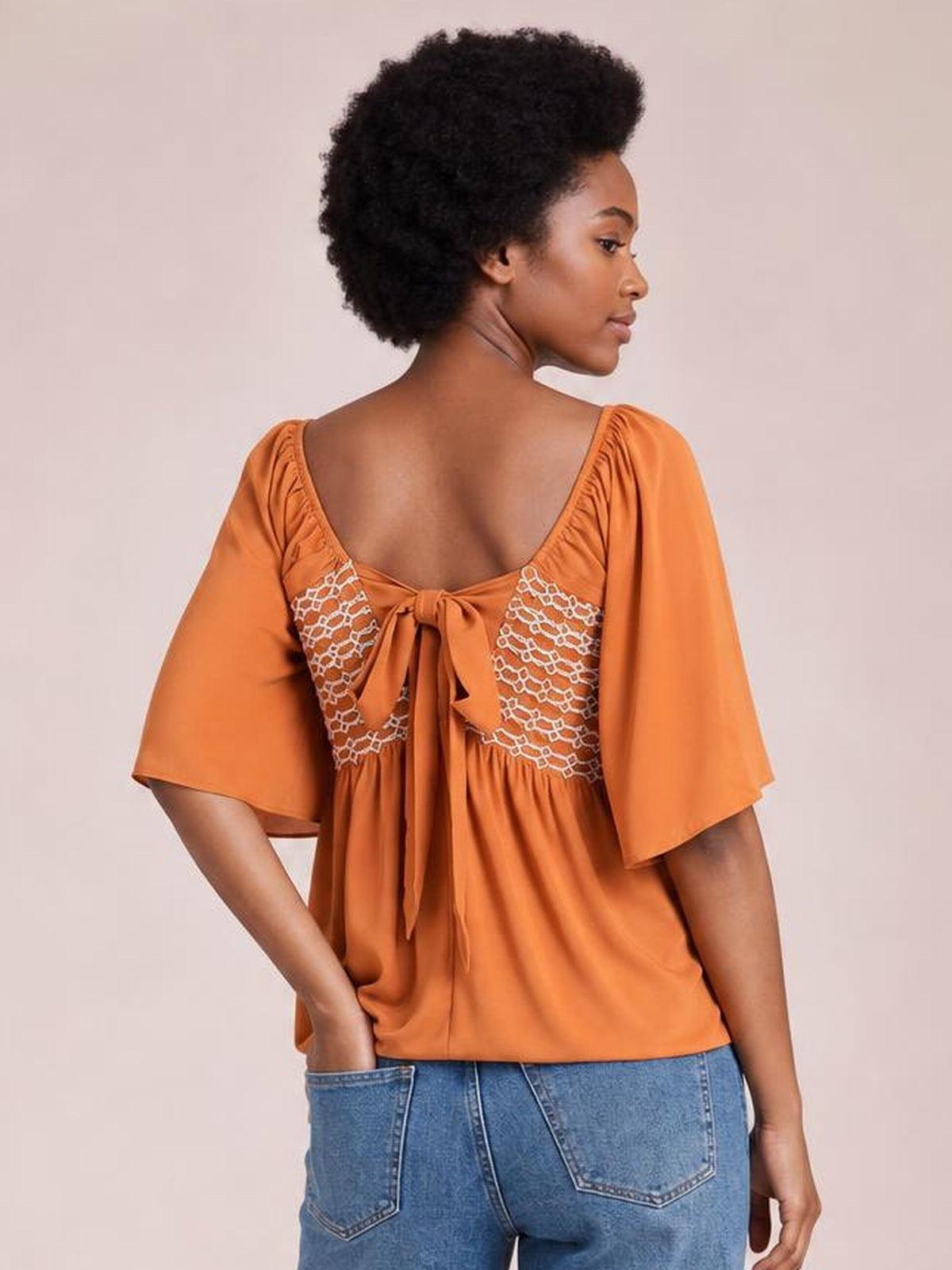 Model shown from behind wearing an orange tied square-neck blouse with half sleeves, a relaxed silhouette, gathered seams, and a back bow detail.