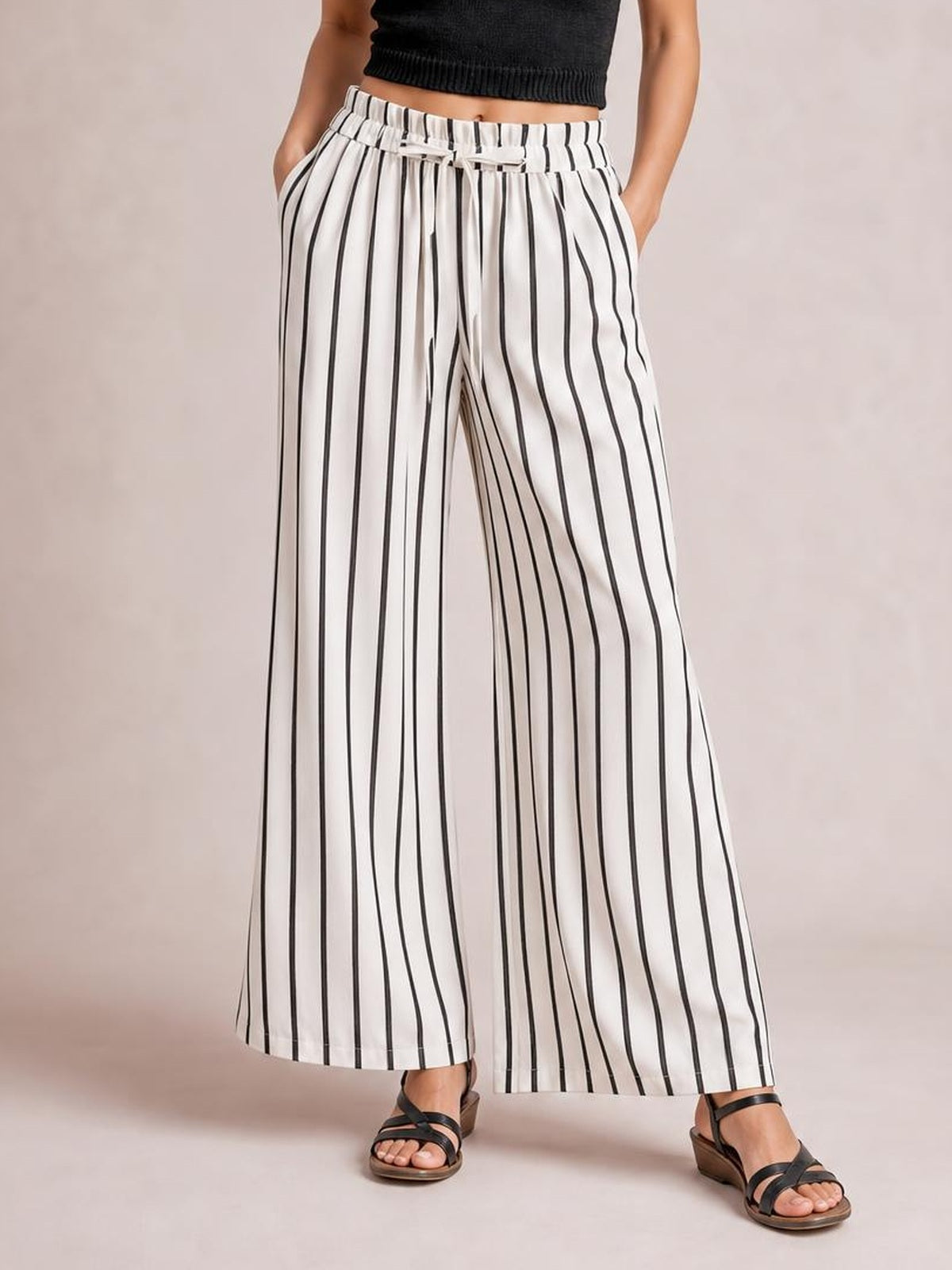 Model poses with hands in the pockets of white opaque pants featuring black vertical stripes, a tied waistband, and a flowy wide-leg silhouette.