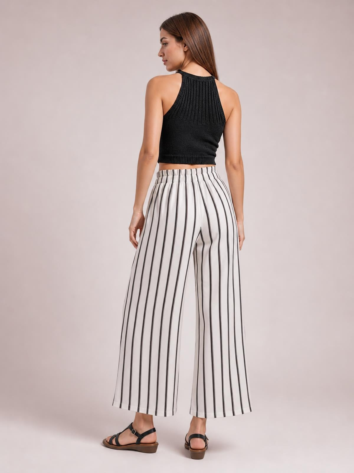 A model stands turned slightly away, showcasing white-and-black vertically striped wide-leg pants with a gathered high waistband and flowing silhouette.