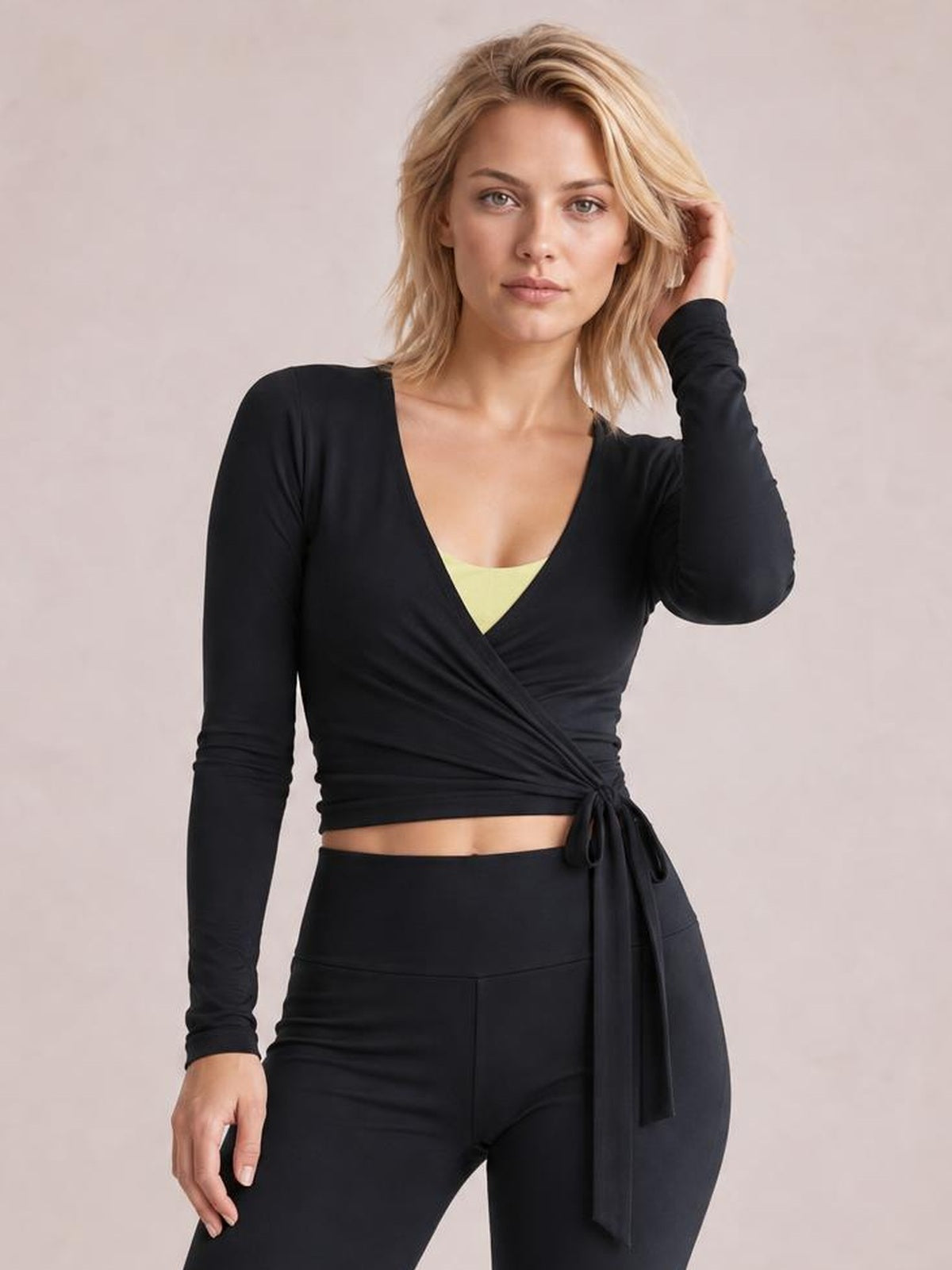A model poses with one hand in her hair, wearing a black long-sleeve surplice top with a wrap silhouette, crossover front, and side waist tie over black pants.