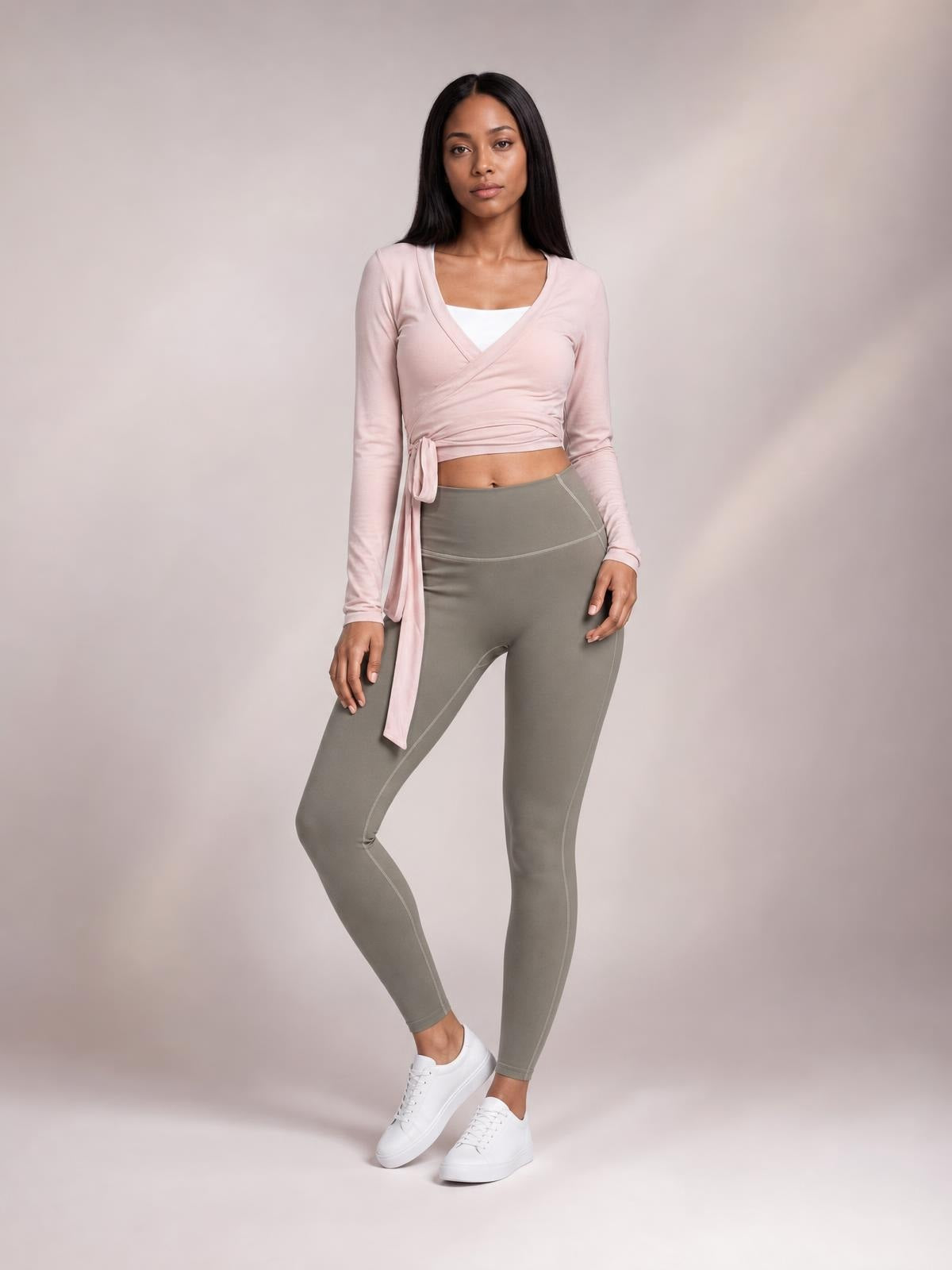 A model stands with one leg bent in a blush pink tied surplice long sleeve top with a wrap front, cropped silhouette, and slim fit.
