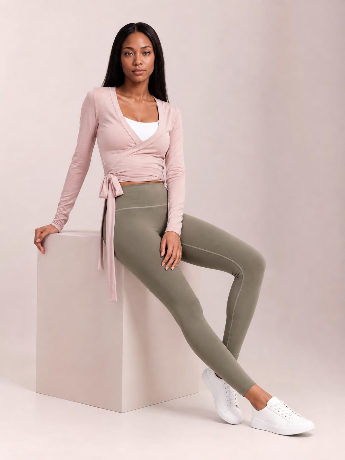 A model seated in a relaxed pose wears a blush pink tied surplice long sleeve top with a crossover neckline, fitted cropped silhouette, and waist sash.