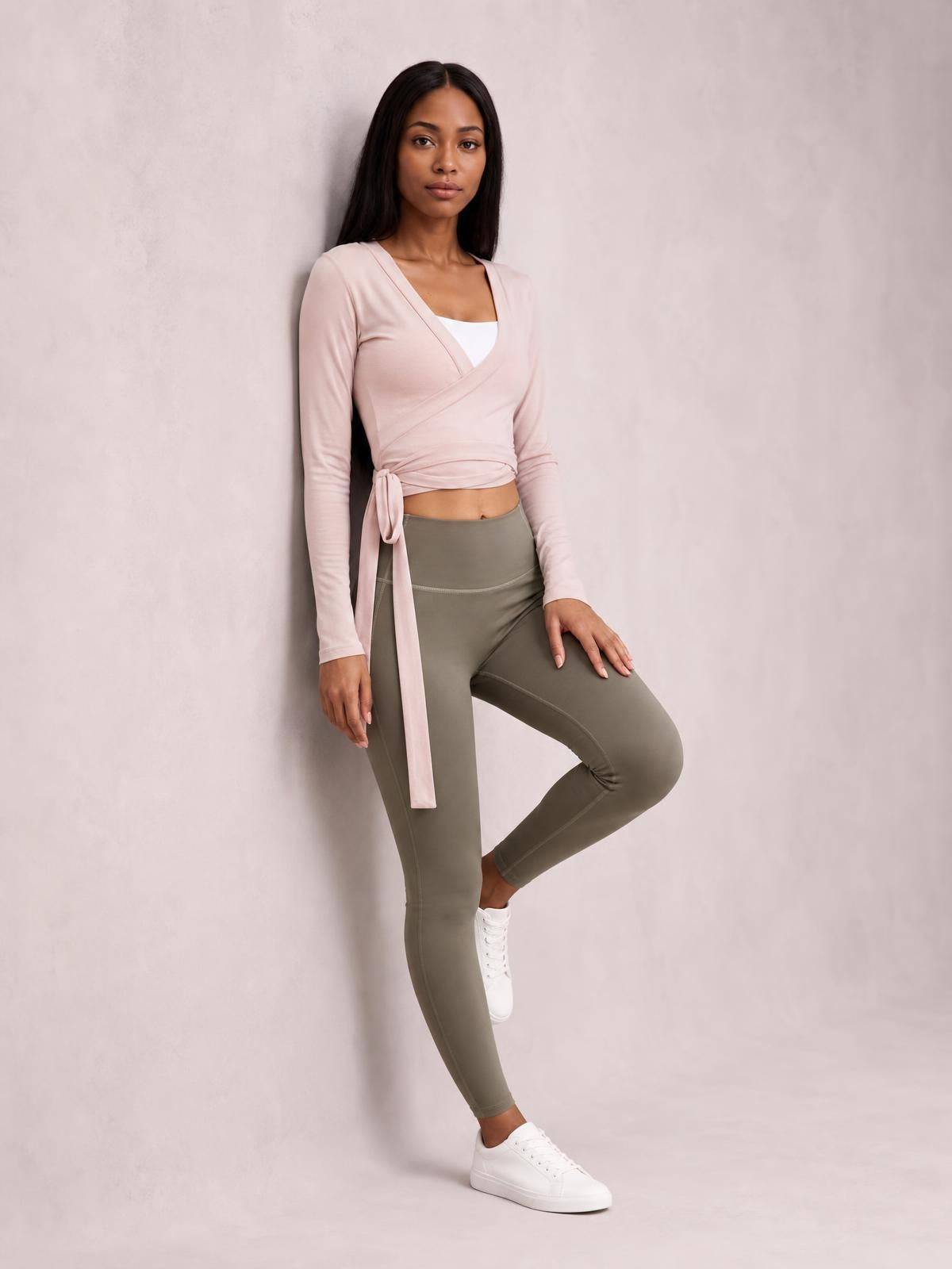 Model wears a blush pink tied surplice long sleeve top with a wrap-style neckline, fitted silhouette, and long waist ties, posing with one leg raised.