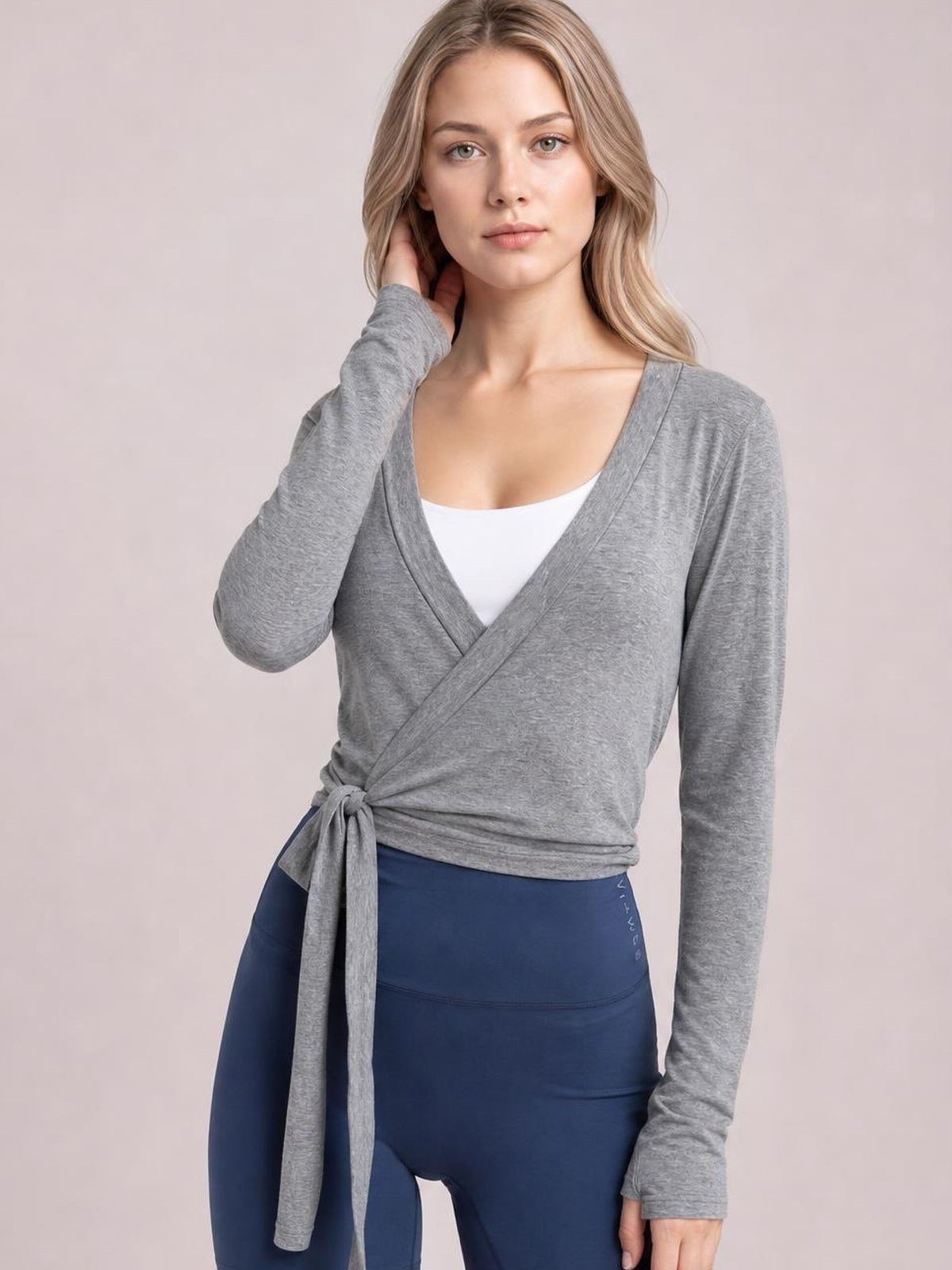 Model poses with one hand at her neck wearing a gray tied surplice long sleeve top with a wrap-front silhouette and waist tie.