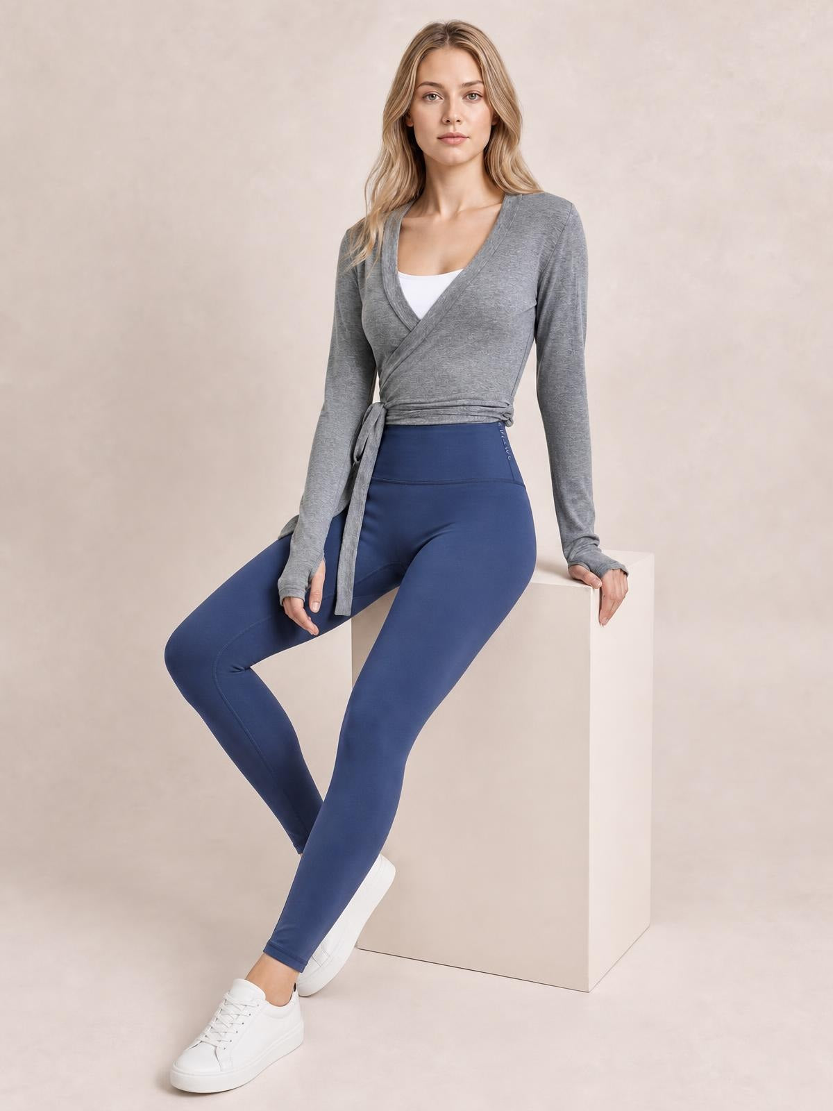 A model sits in a gray tied surplice long sleeve top with a wrap front, waist sash, fitted silhouette, and white layer visible at the neckline.