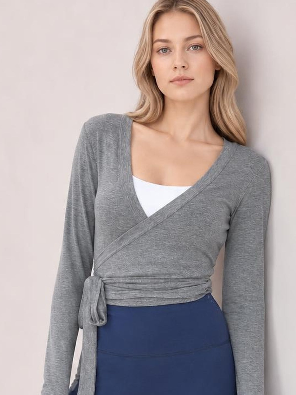 Model wears a gray tied surplice long sleeve top with a wrap-style front, waist sash, and fitted silhouette over a white camisole.