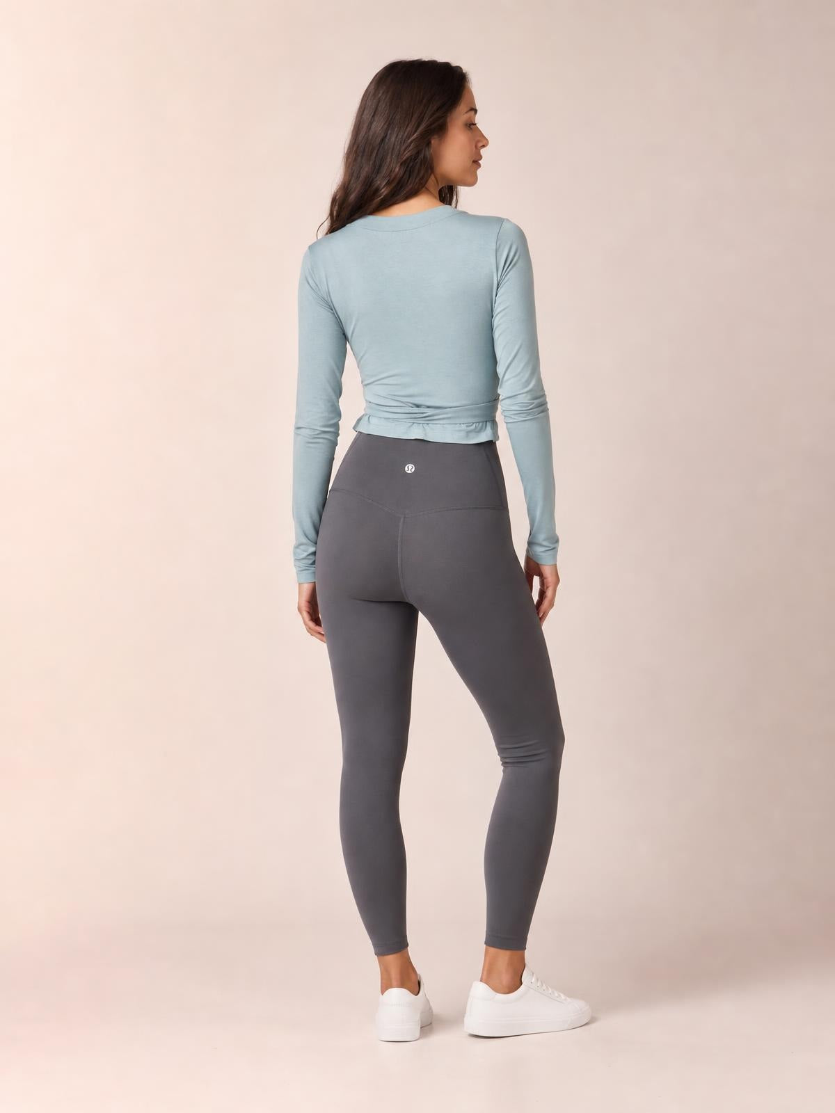 A model poses in a back view wearing a pale blue fitted Tied Surplice Long Sleeve Top with a cinched, tied waist and long sleeves.