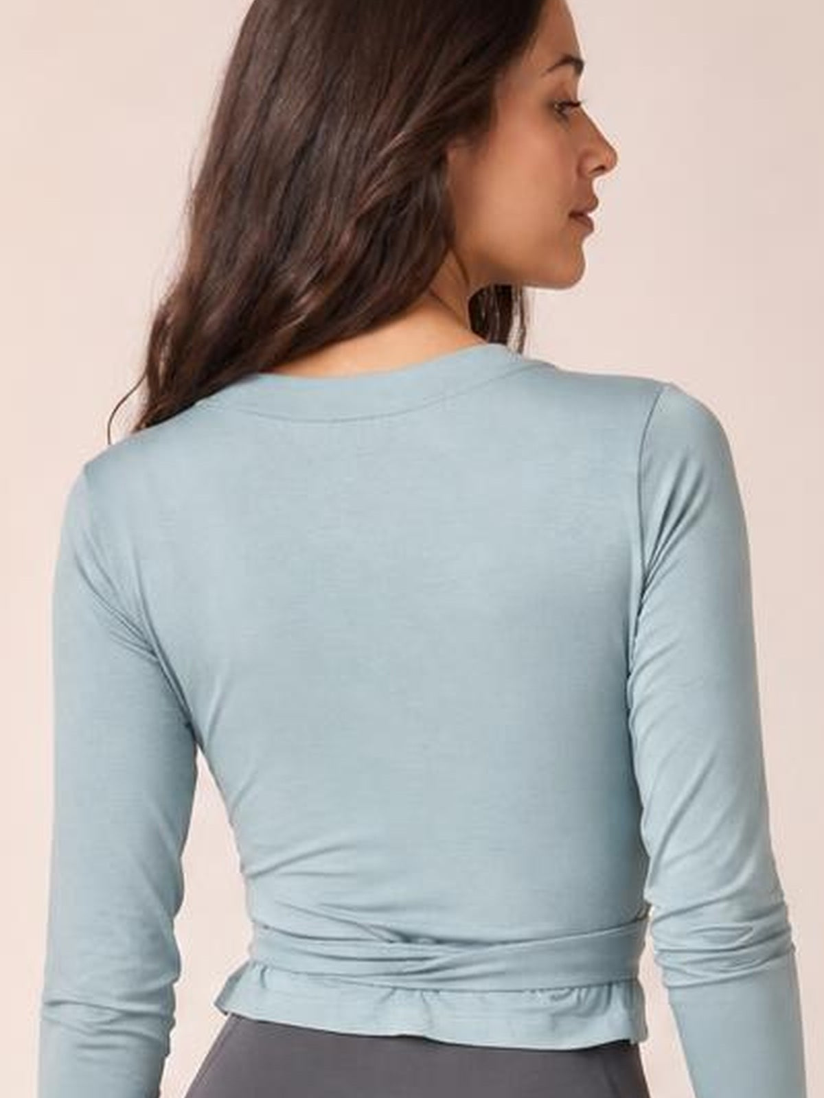 A model poses in profile wearing a light blue tied surplice top with long sleeves, a fitted silhouette, wrap construction, and a peplum-like hem.