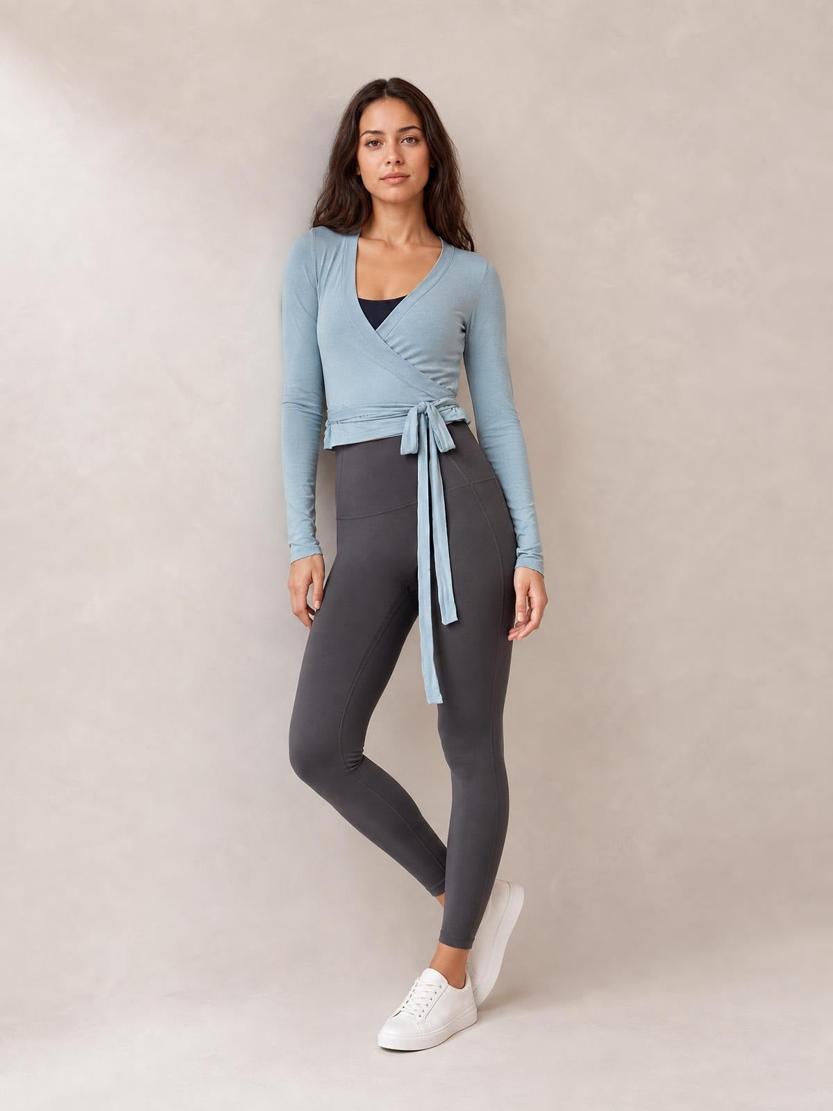 Model poses in a light blue tied surplice long sleeve top with a fitted cropped silhouette, wrap-front construction, and long waist ties.