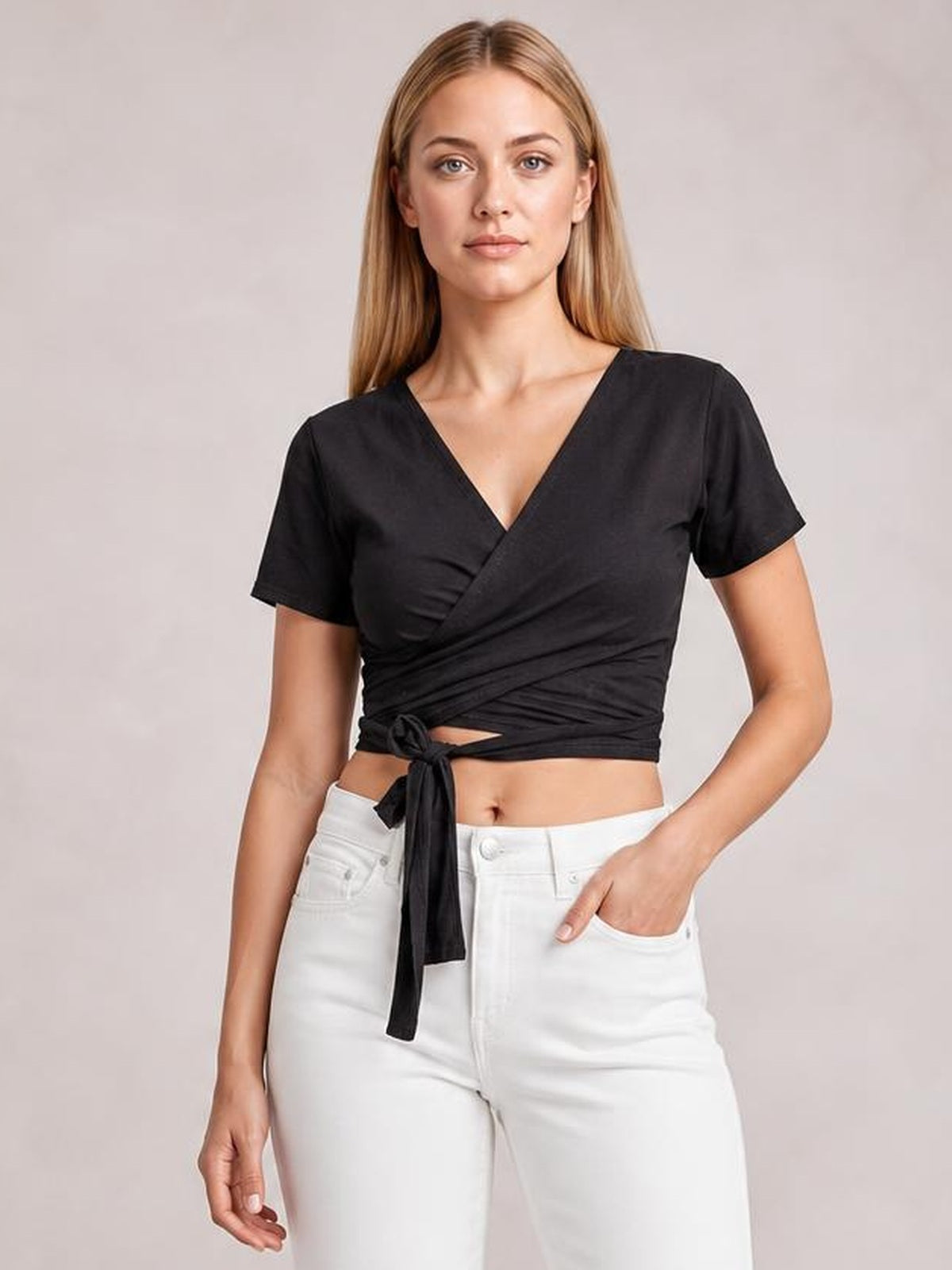 Model wearing a black tied surplice short sleeve top with a V-neck wrap silhouette, cropped hem, and front waist tie, posing with one hand in her pocket.
