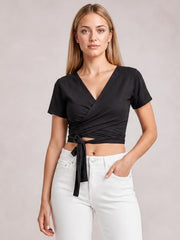 Model wearing a black tied surplice short sleeve top with a V-neck wrap silhouette, cropped hem, and front waist tie, posing with one hand in her pocket.
