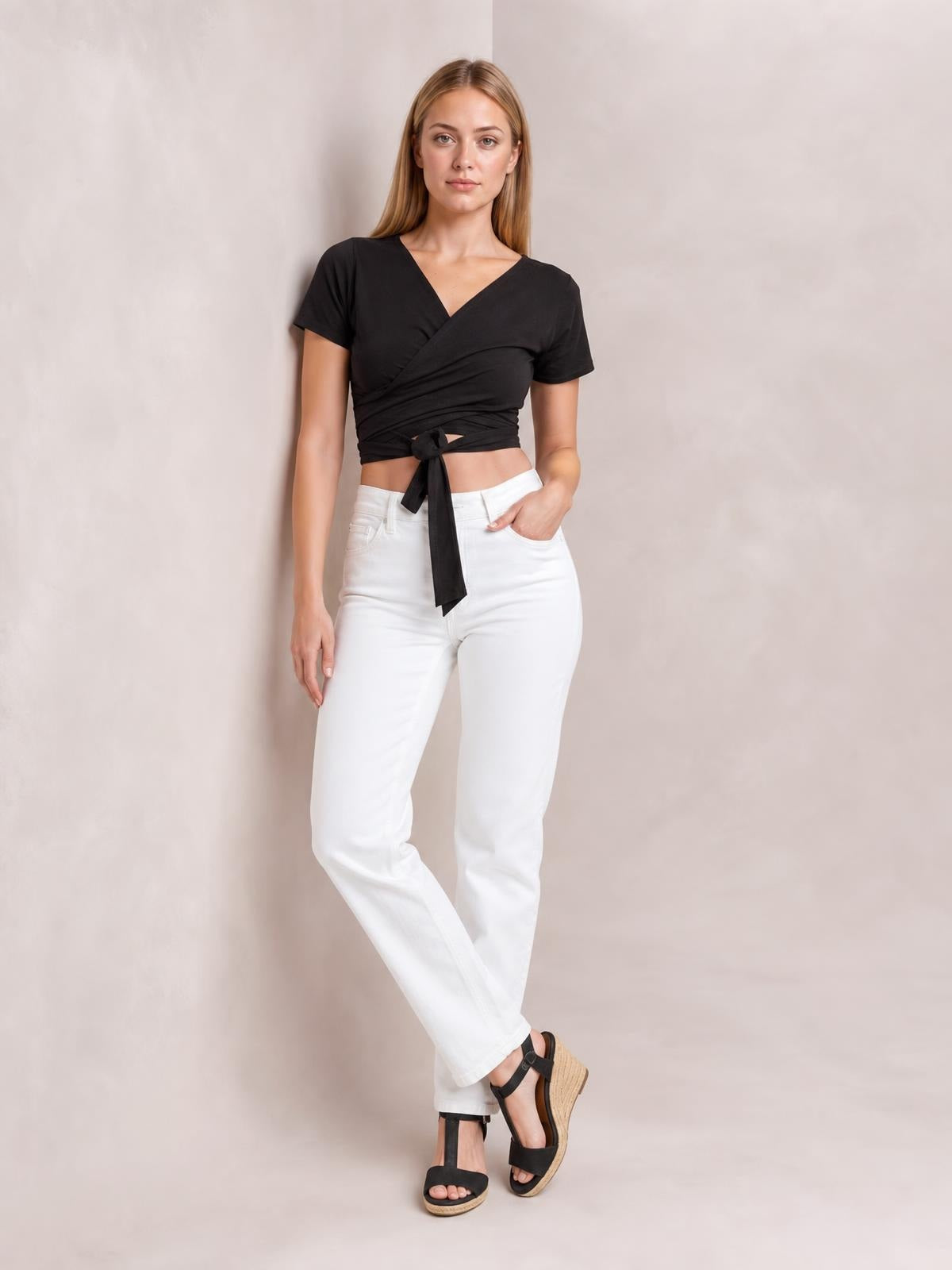 Model in a black tied surplice short sleeve top with a fitted cropped silhouette, standing with crossed legs and one hand in her pocket.