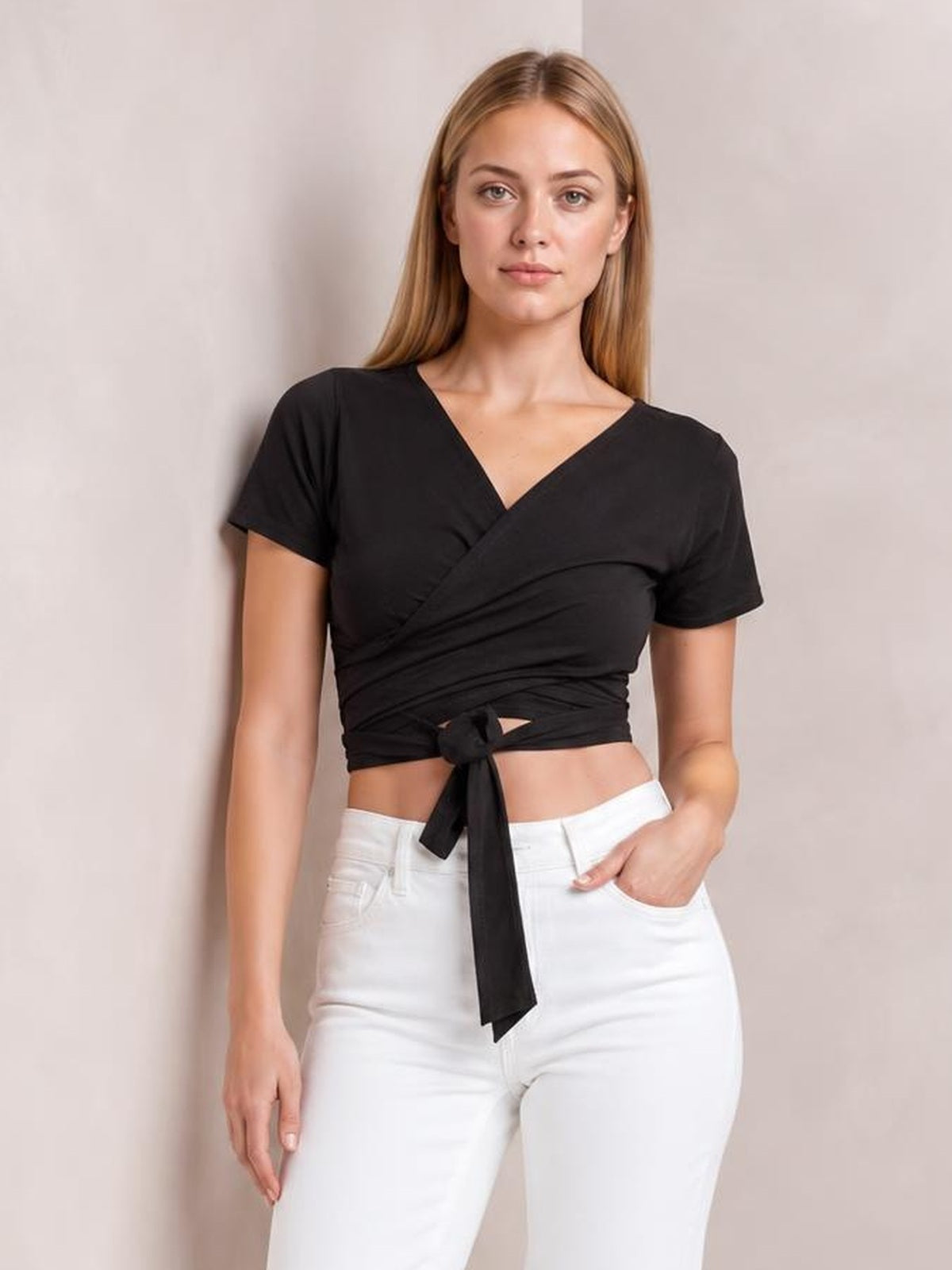 Model poses in a black tied surplice short sleeve top with a V-neck, wrapped bodice, fitted cropped silhouette, and long waist tie.
