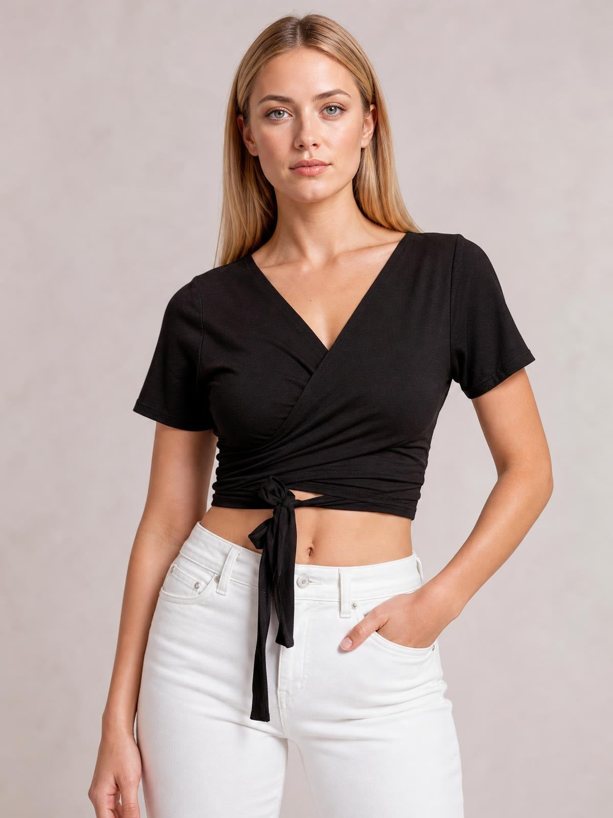A model poses with one hand in her pocket, wearing a black tied surplice short sleeve top with a V-neck, wrap construction, and cropped silhouette.