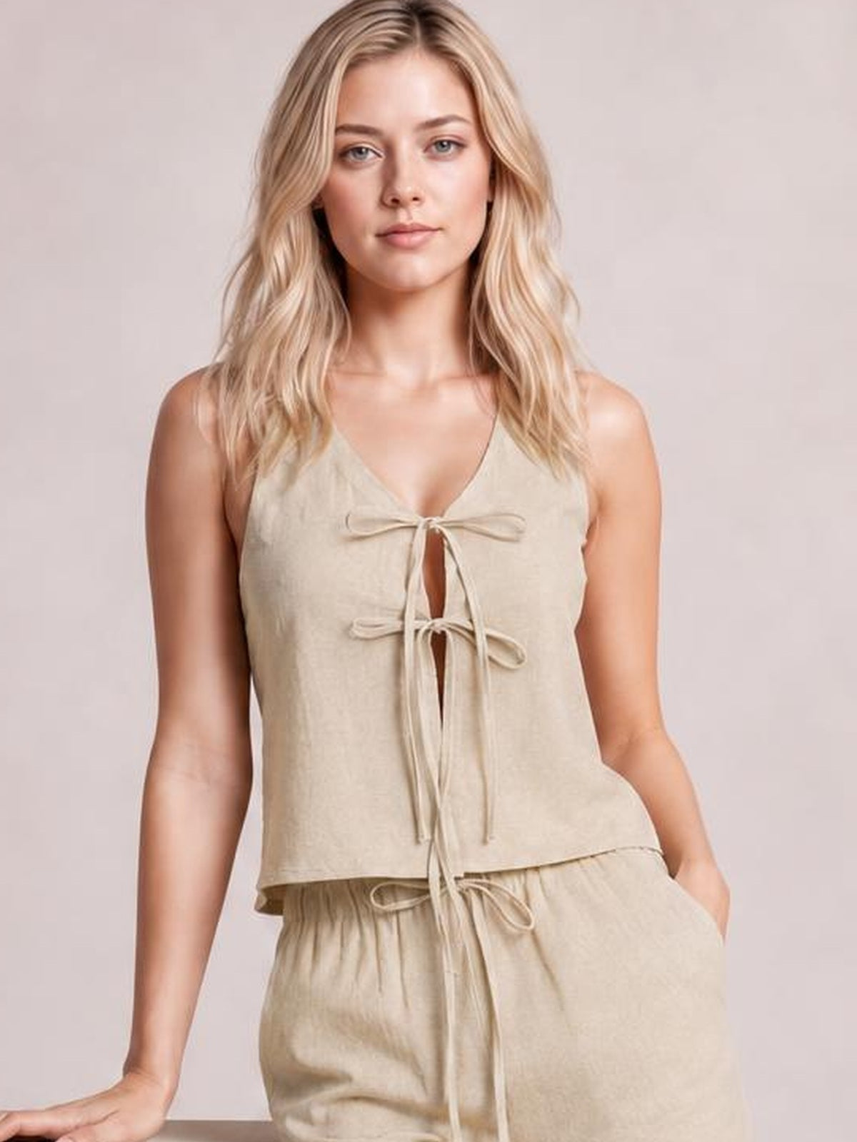 Model wearing a beige sleeveless V-neck top and matching drawstring pants set, with tied front detail, relaxed silhouette, and a hand in a pocket.