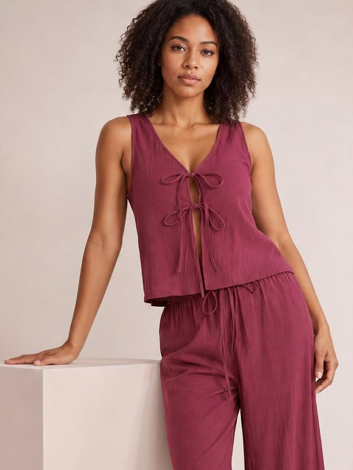 A model leans against a cube wearing a burgundy tied V-neck sleeveless top with a relaxed silhouette and matching drawstring wide-leg pants.