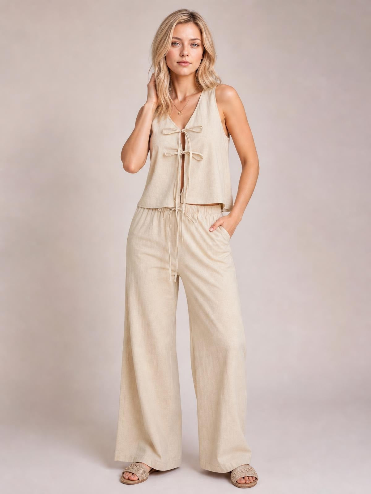 A model poses in a beige tied V-neck sleeveless top and matching wide-leg pants with a drawstring waist and pockets, one hand in a pocket.