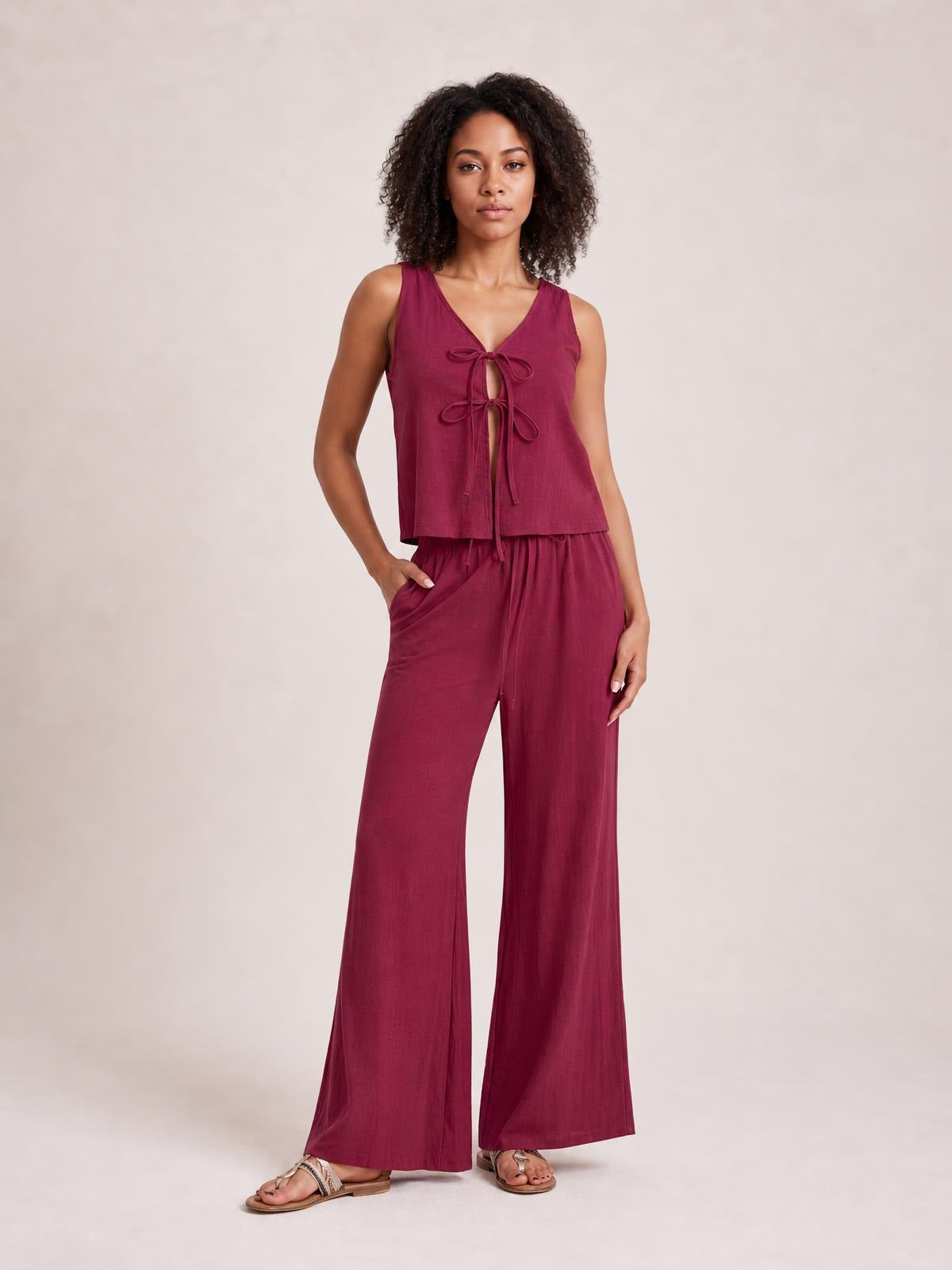 Model poses in a burgundy tied V-neck sleeveless top with front cutout details and matching wide-leg drawstring pants with pockets.