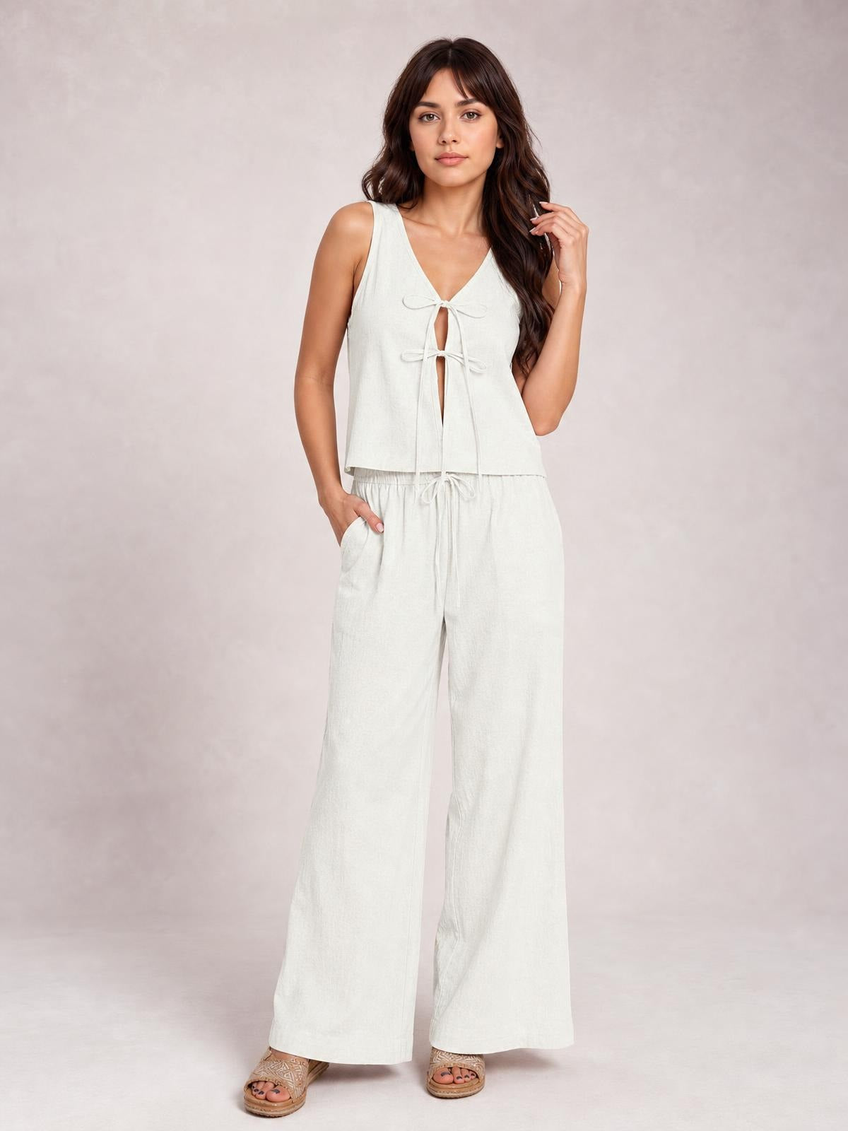 Model wears an ivory sleeveless V-neck top with front tie details and matching wide-leg drawstring pants with pockets, posing with one hand in a pocket.