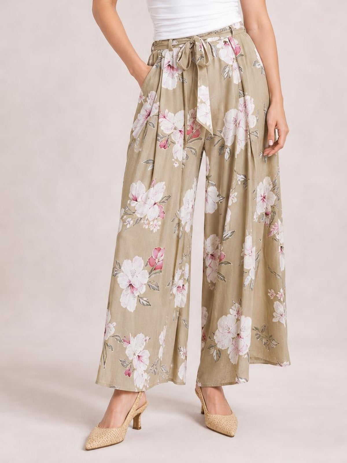 A model wears tan floral wide-leg pants with a tied waist, flowing silhouette, and large white and pink blossoms, posing with one hand in a pocket.