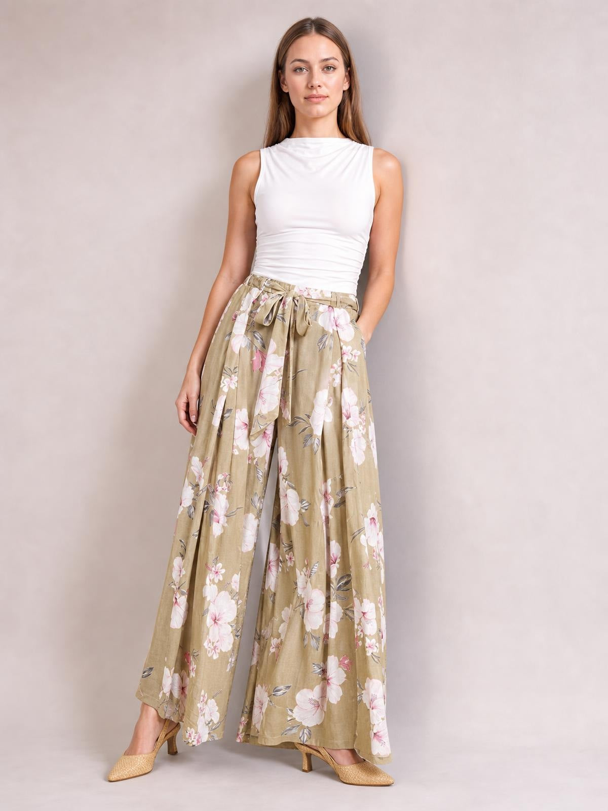 A model poses in tan floral wide-leg pants with a tied waist and flowing slit-leg construction, styled with a fitted white sleeveless top.