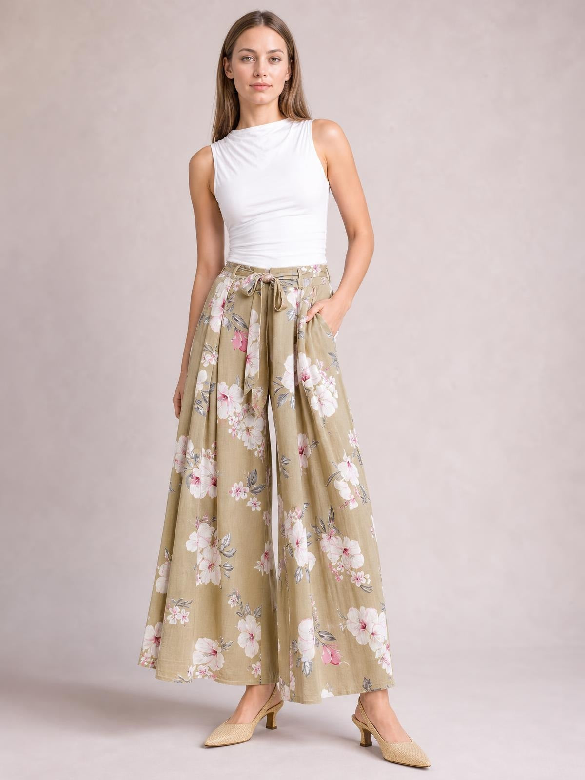 A model poses in beige floral wide-leg pants with a tied high waist, flowing silhouette, and visible side pockets, styled with a white sleeveless top.