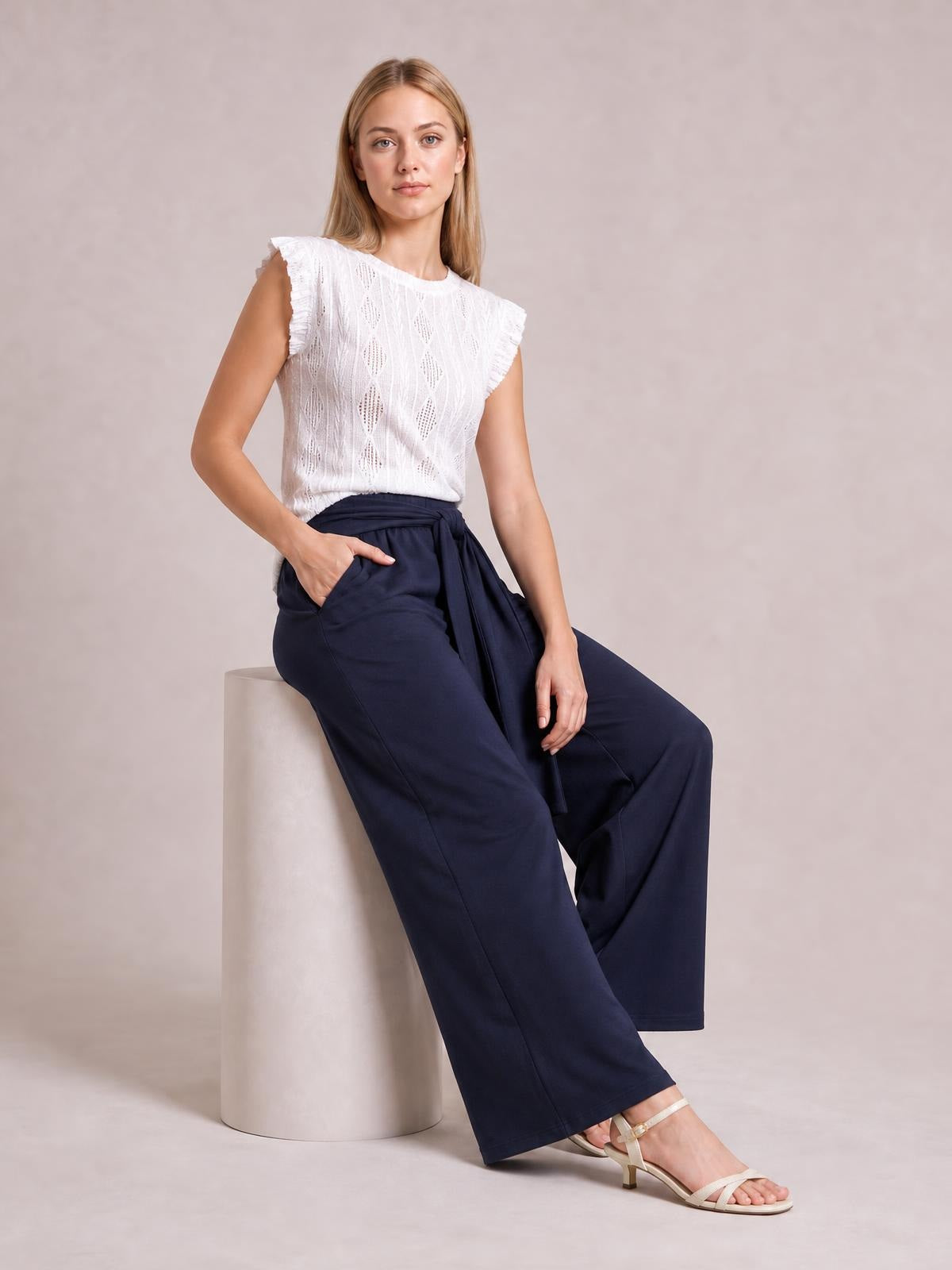 Seated model wears navy tied wide leg pants with a draped waist sash, visible side pockets, and a relaxed full-length silhouette.