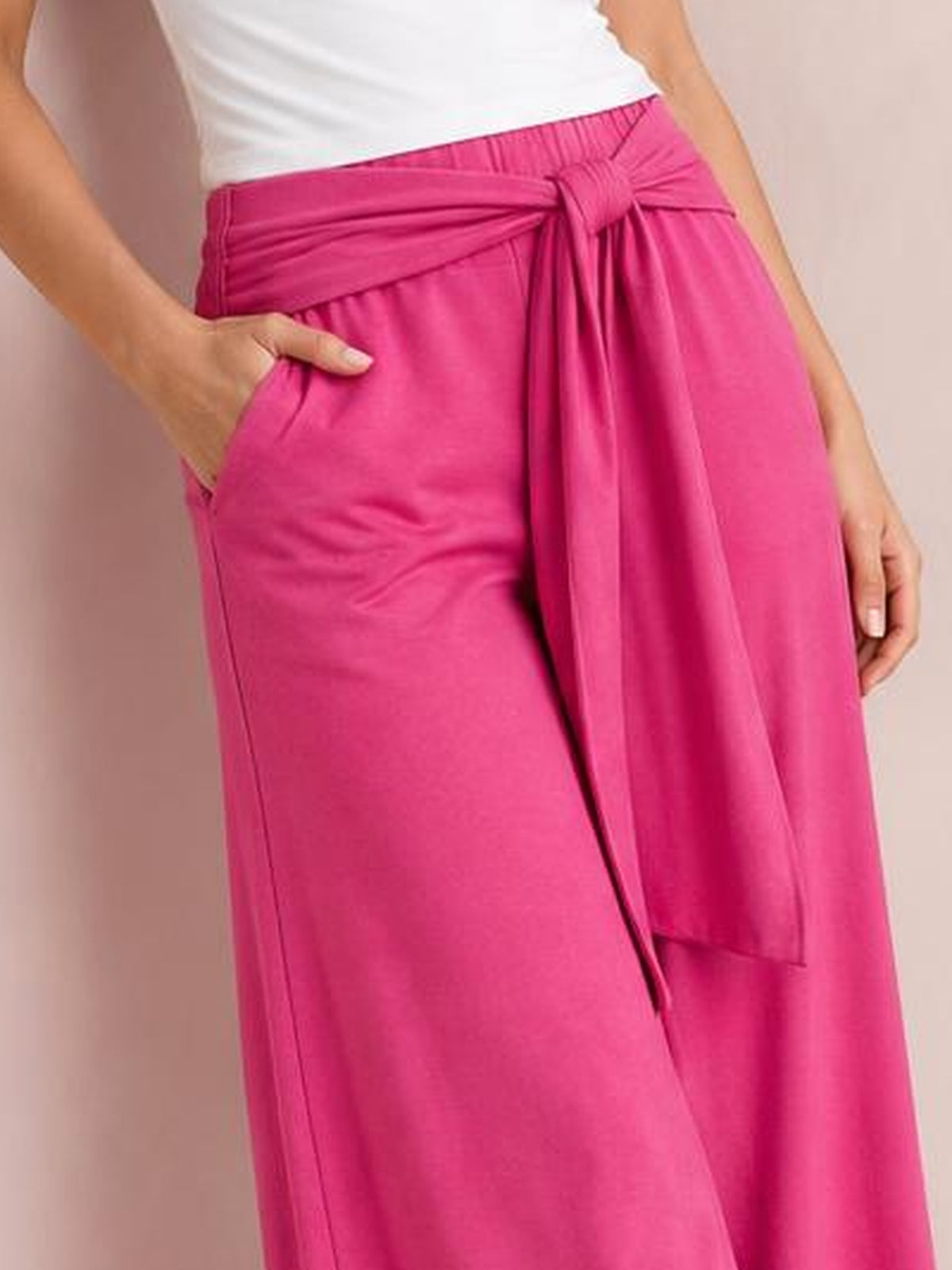 Model poses with one hand in a pocket, showing bright pink tied wide-leg pants with a gathered waistband and long waist sash.