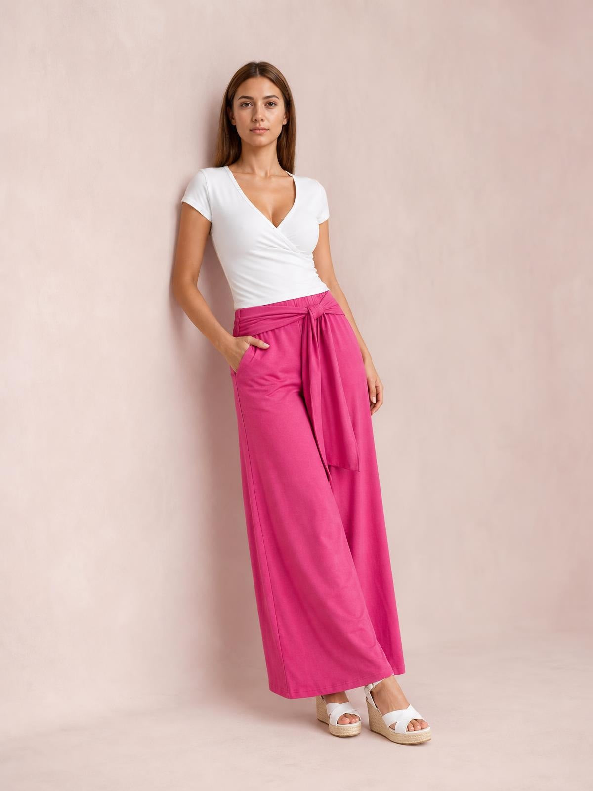 Model poses in bright pink tied wide-leg pants with side pockets, a flowing full-length silhouette, and a tied waist detail.