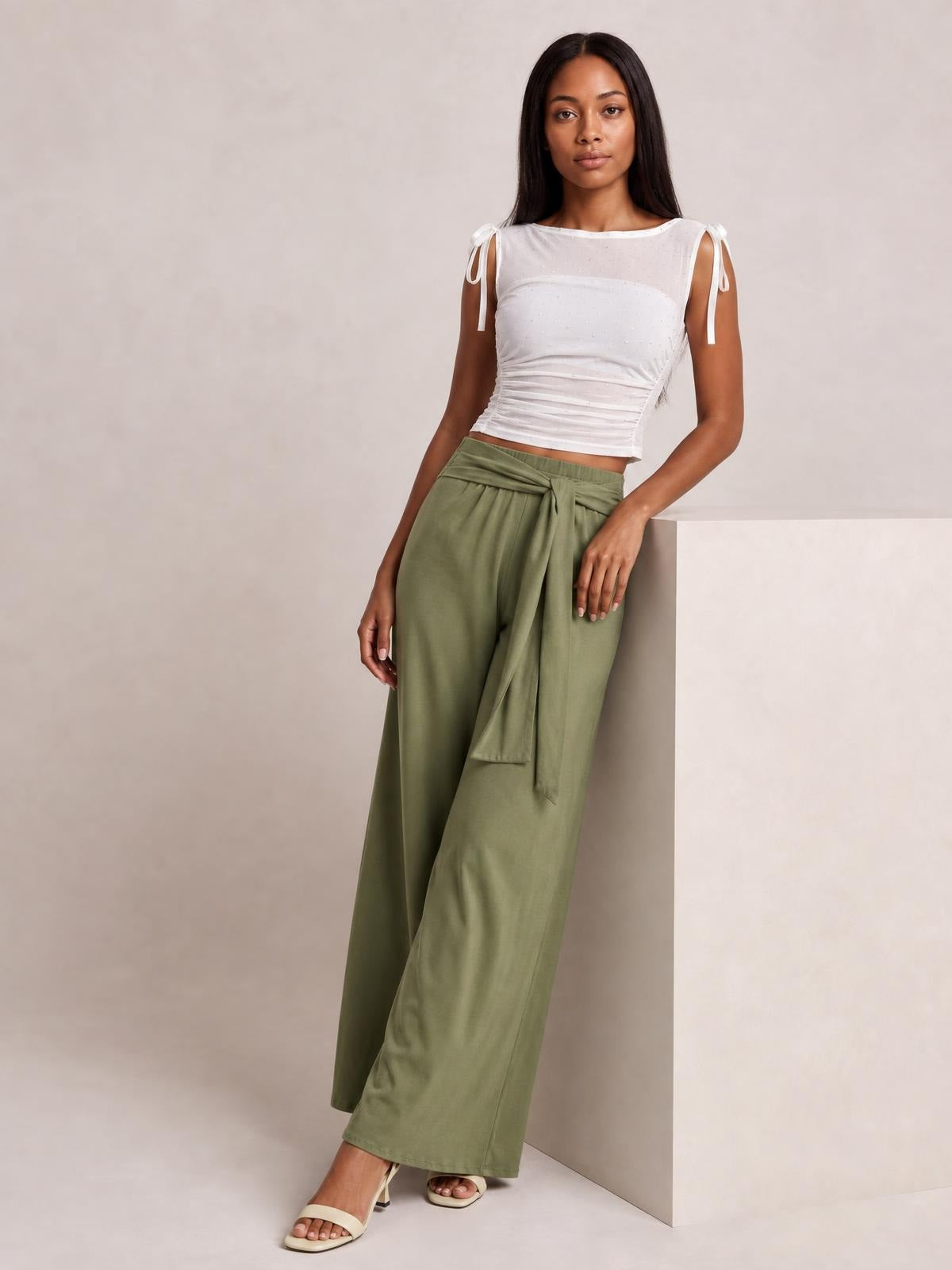 Model leans against a wall wearing olive green tied wide leg pants with a flowing silhouette, high waist, front tie, and visible pockets.