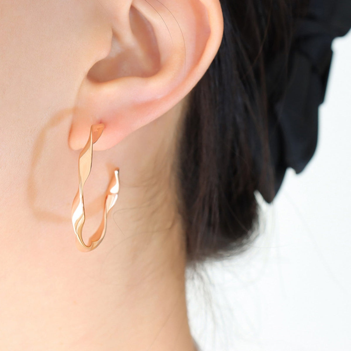 Close-up of a model wearing polished gold-plated titanium steel hoop earrings with an elongated, gently twisted open silhouette.
