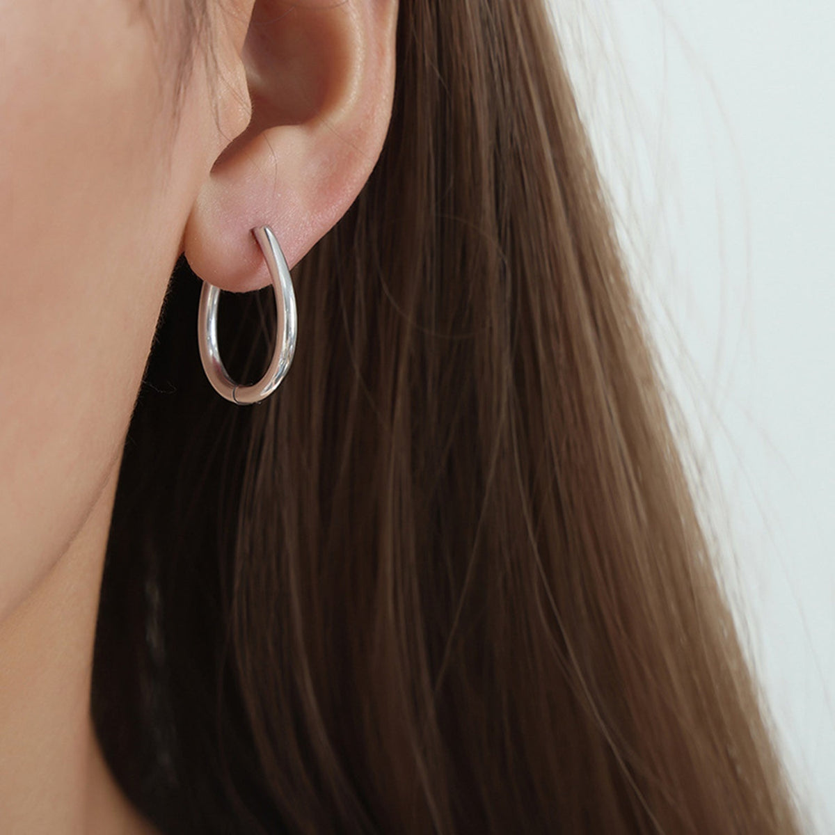 A close-up shows a polished silver-tone titanium steel huggie earring with a compact oval hoop silhouette worn on the model’s ear.