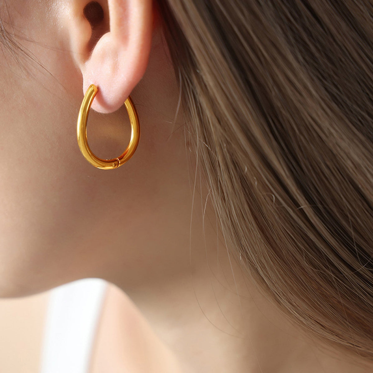 Close-up of a model wearing a polished gold-tone Titanium Steel Huggie Earring with a compact rounded hoop silhouette and hinged closure.