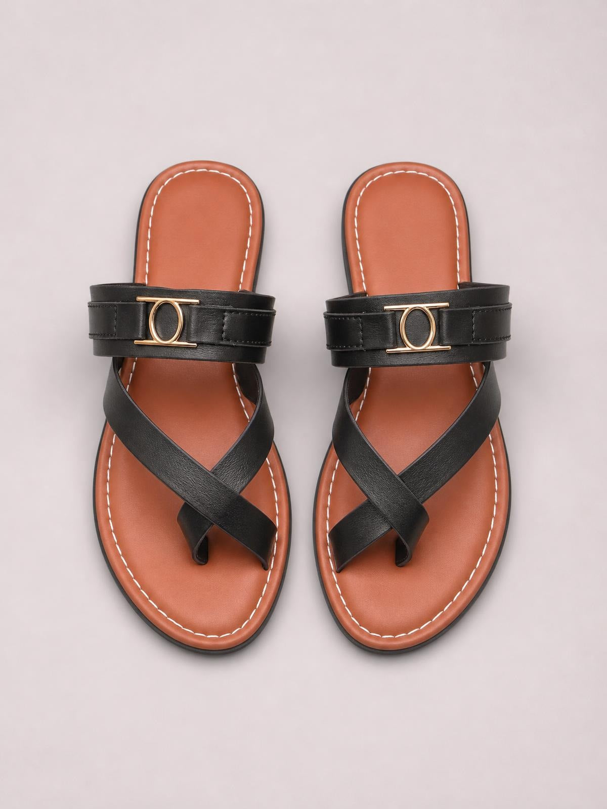 Black toe loop flat sandals feature crisscross straps, a wide gold-tone circular ornament, tan cushioned footbeds, and contrast stitching.