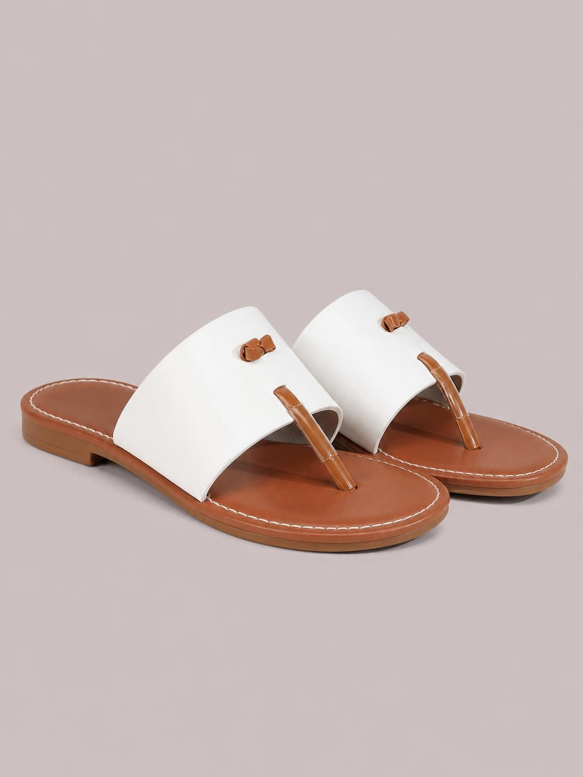 White toe post flat sandals feature wide PU leather straps, brown rubber footbeds, bow accents, and stitched edging.