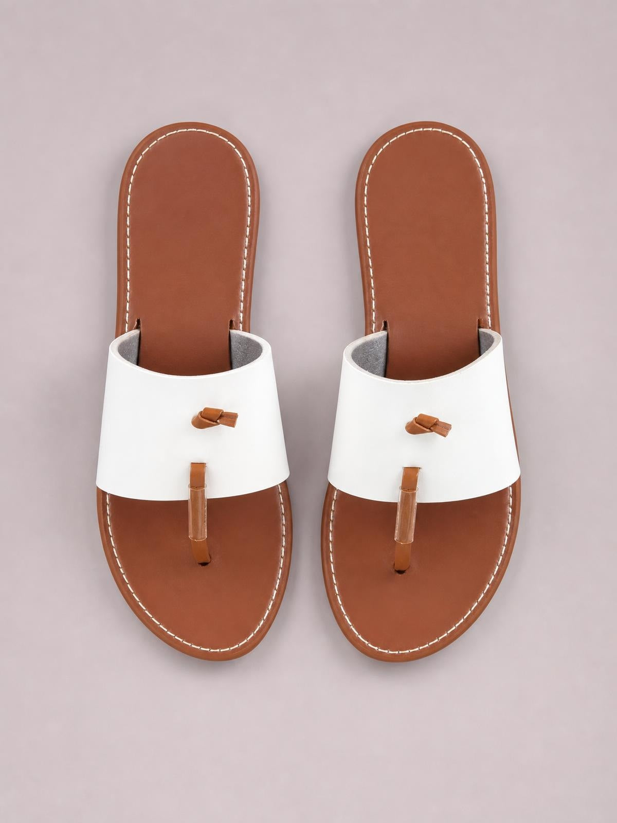 White and brown toe post flat sandals feature wide upper straps, rounded open toes, tan soles, and contrasting stitched construction.
